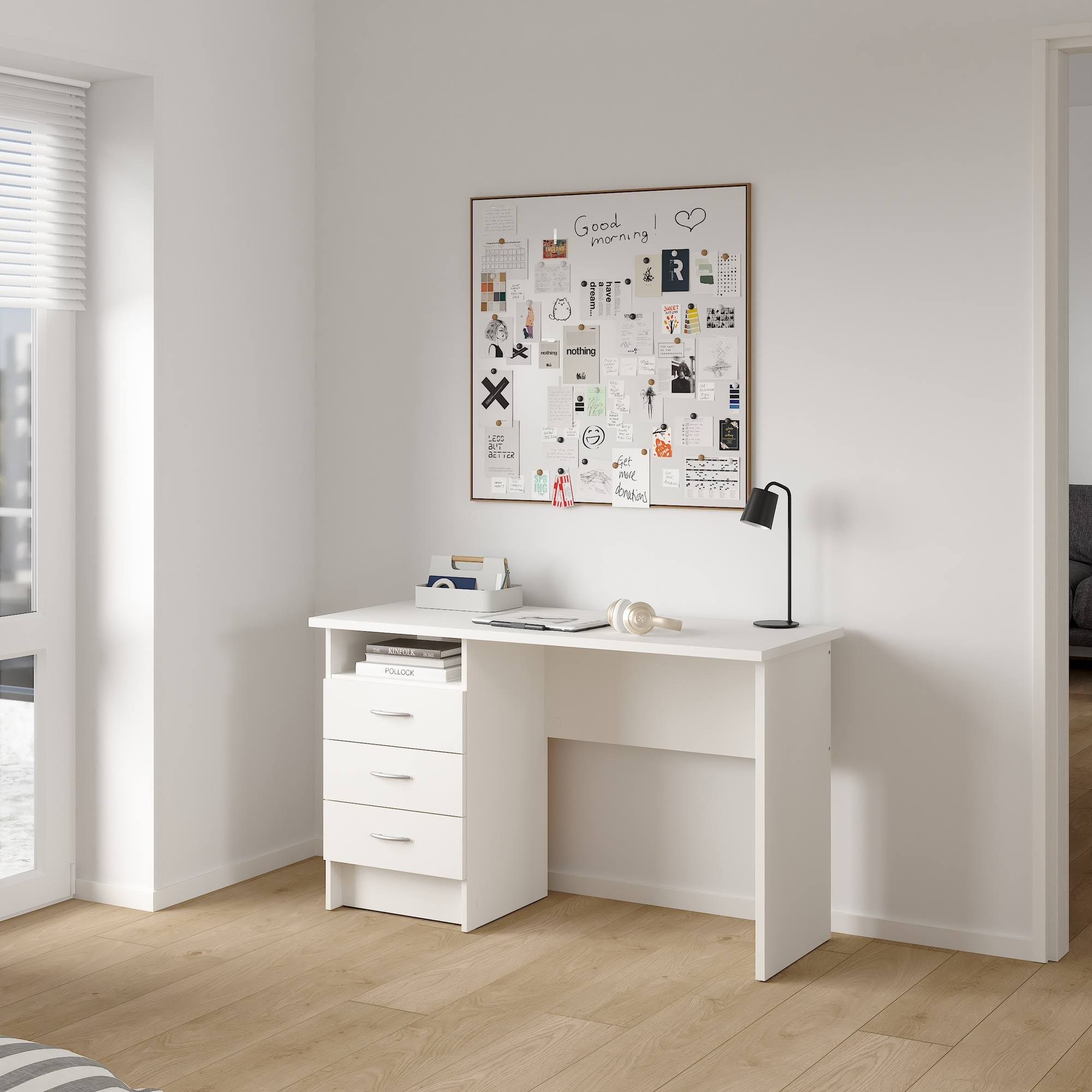 Un escritorio blanco con cajones, una pizarra con notas, una lámpara negra y auriculares. La habitación tiene pisos de madera.