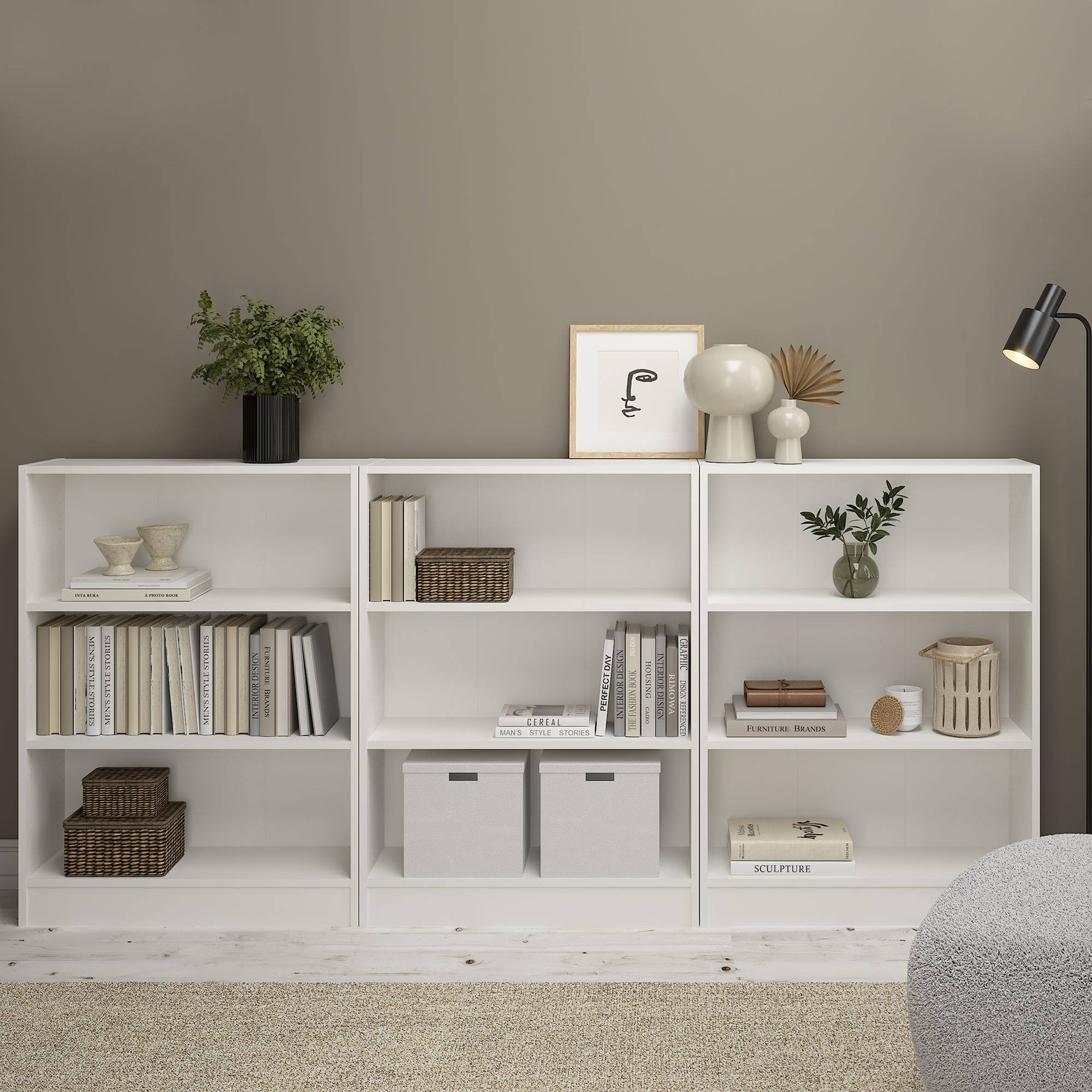 Estanterías blancas con libros, decoración y plantas. Una lámpara y un taburete peludo están al lado. Pared de tonos neutros.