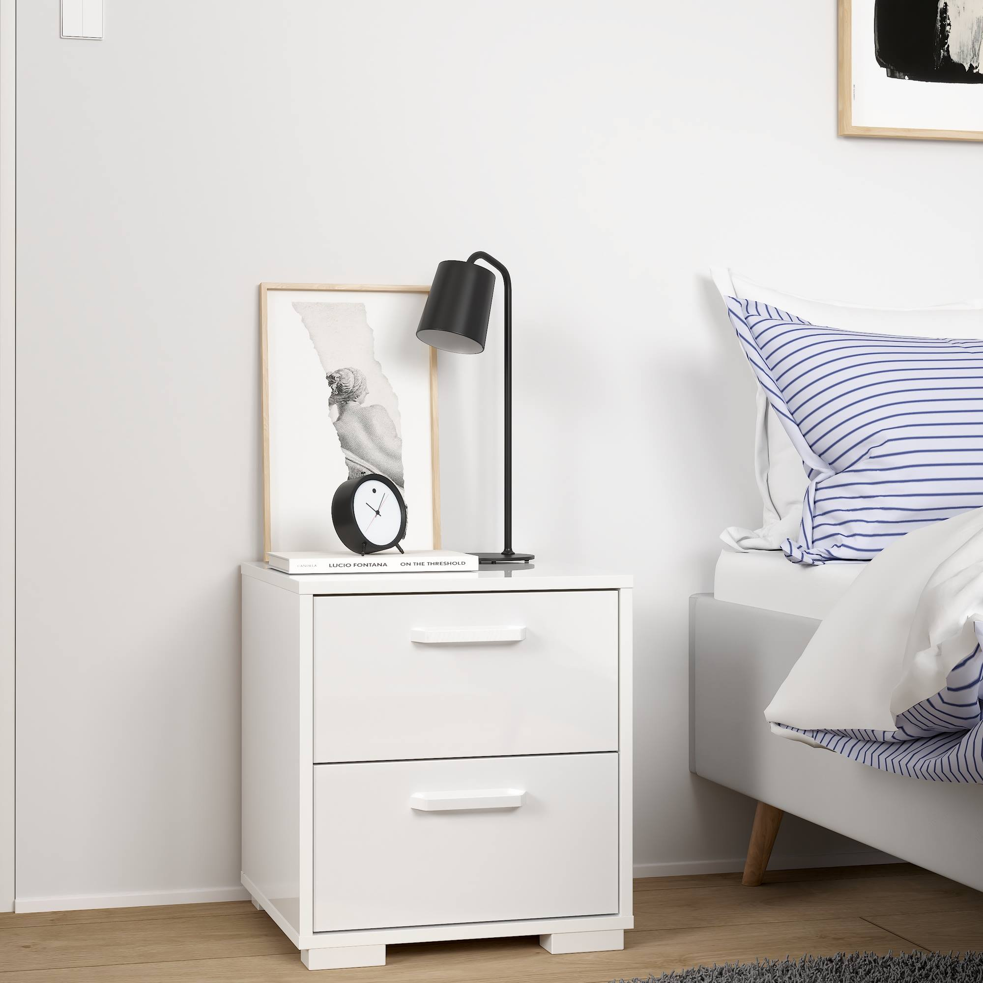 Una mesita de noche blanca con lámpara, reloj y marco. A la derecha, una cama con almohada a rayas. El suelo es de madera.