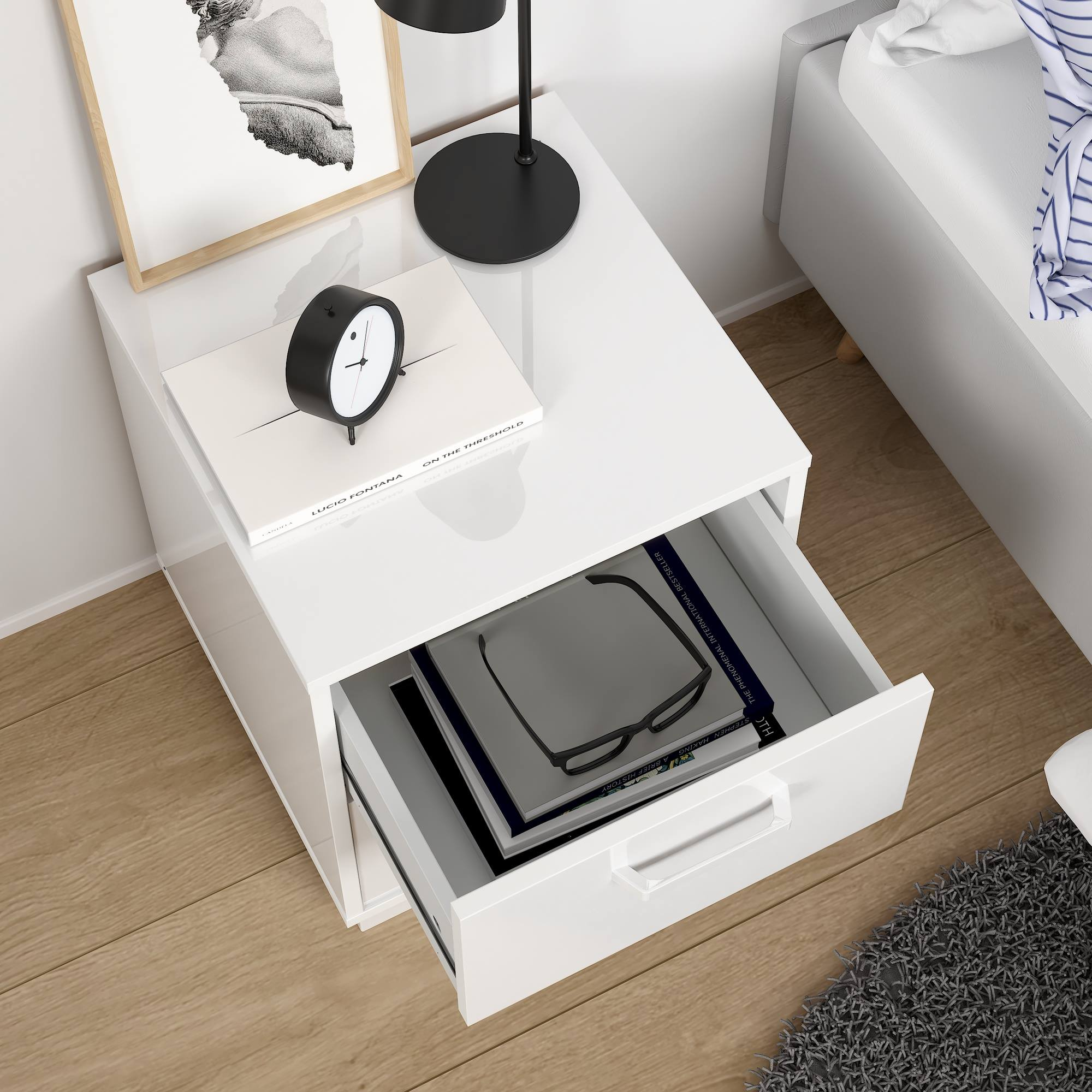 Una mesita de noche blanca con un cajón abierto, libros, gafas y un reloj. Un cuadro y una cama al fondo.