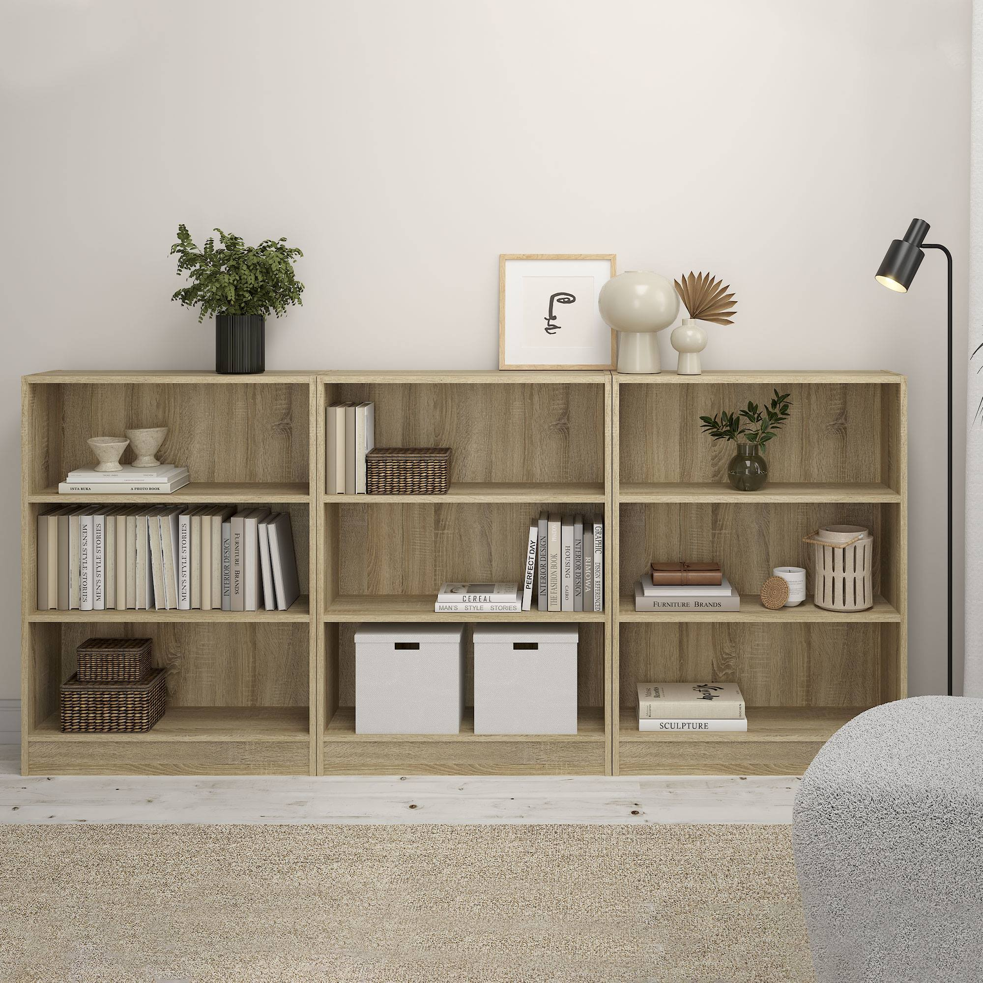 Una estantería de madera con libros, plantas y decoración. Una lámpara negra a la derecha y una silla gris al fondo.