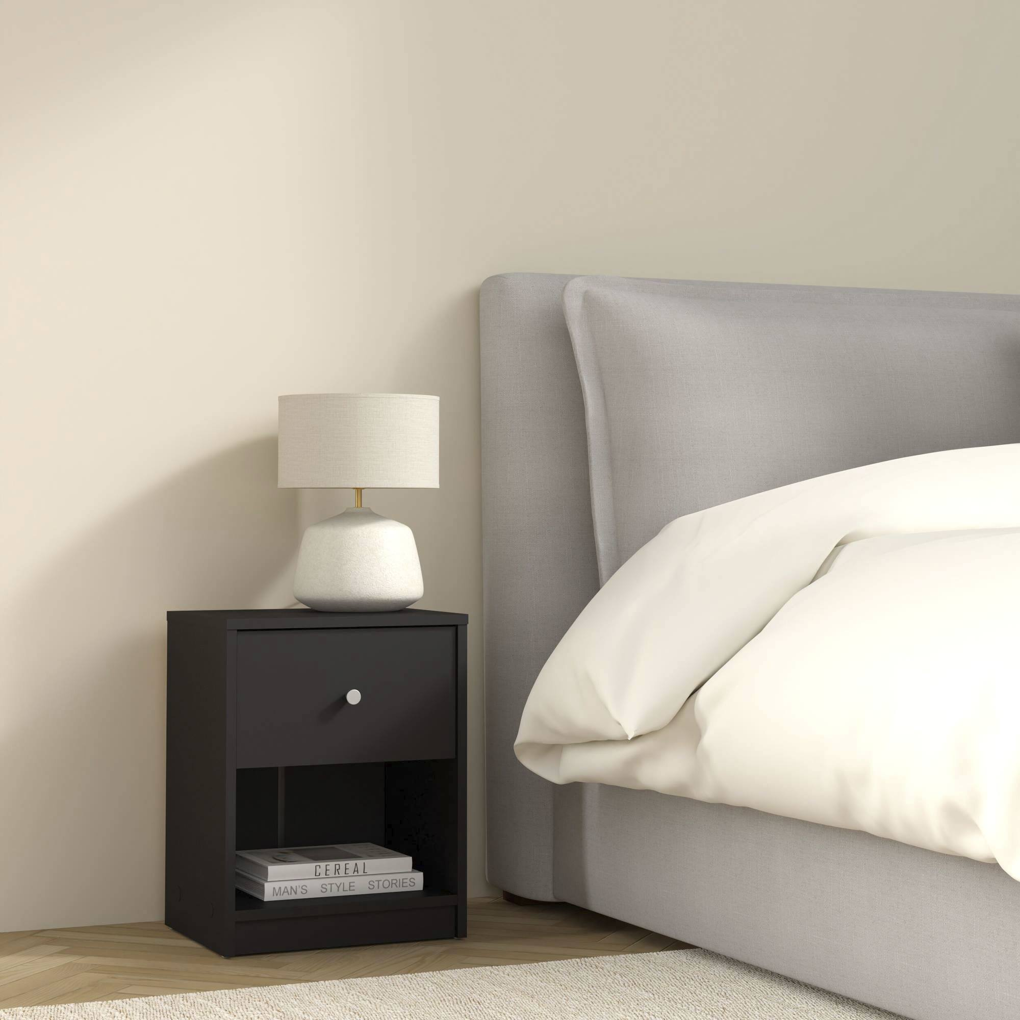 Una escena de dormitorio con una mesita de noche negra, una lámpara y una cama. Libros en el estante inferior. Cama con sábanas blancas.