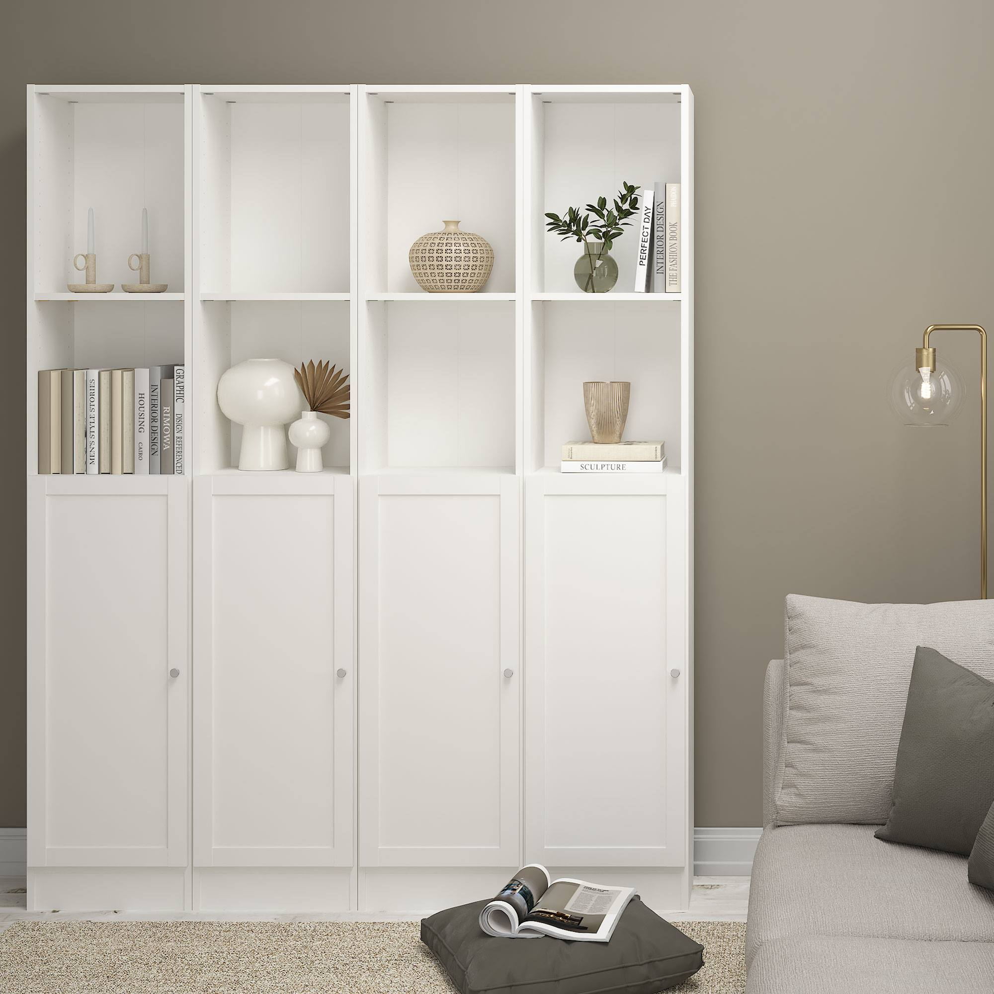 Librería blanca con decoración, libros y un cojín gris con una revista. Un sofá y una lámpara están cerca.