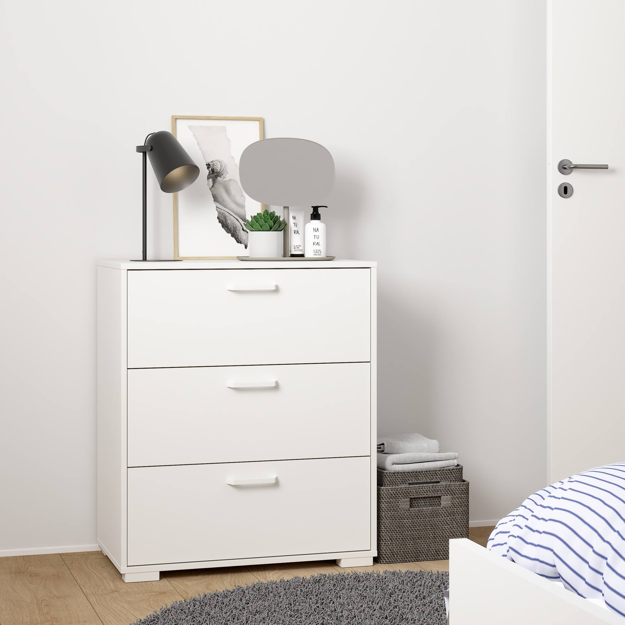 Cómoda blanca con tres cajones, una lámpara, una imagen y una planta. Se ve una cesta y una cama.