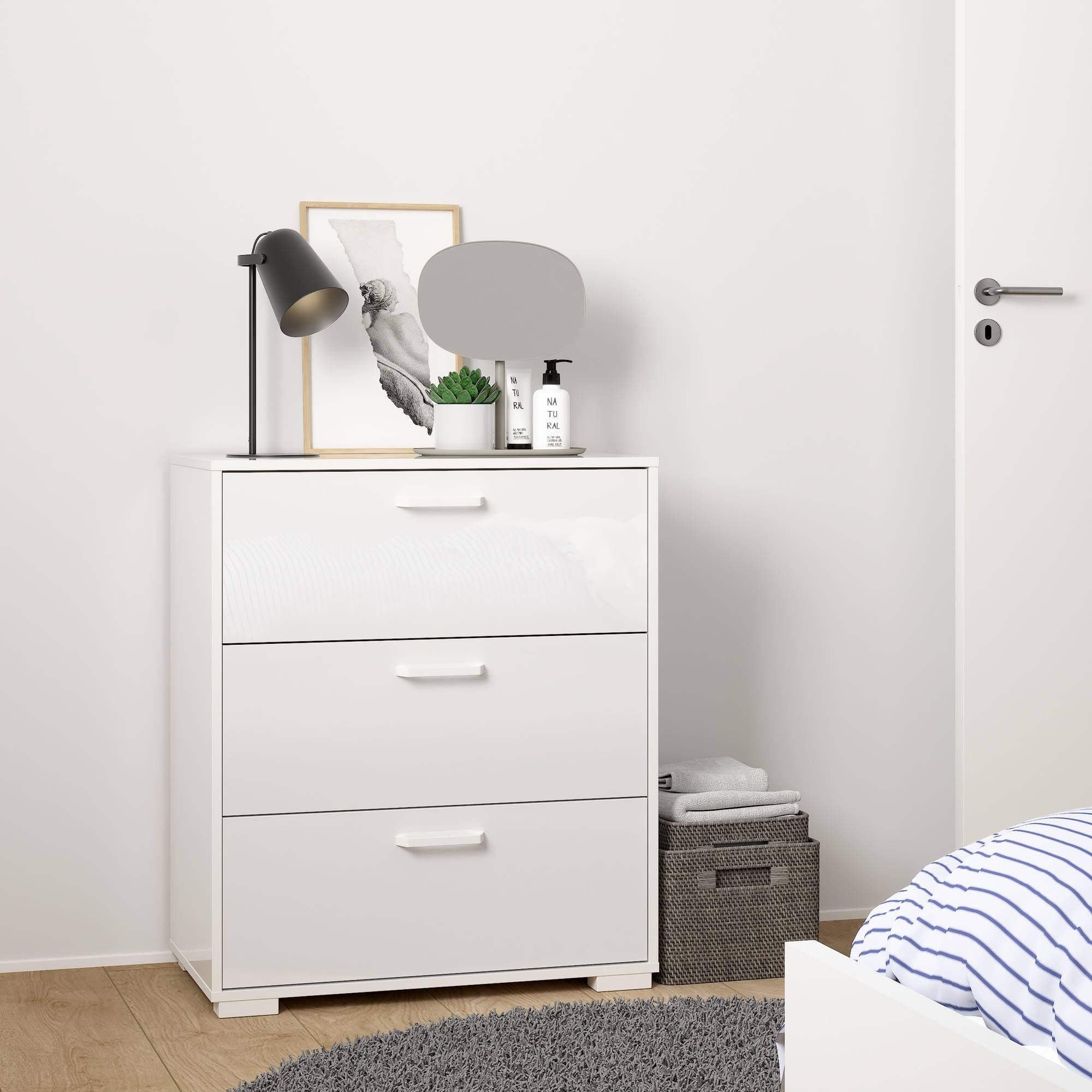 Cómoda blanca con lámpara, arte y planta. Alfombra gris, cesta con toallas y parte de una cama con ropa de cama a rayas.