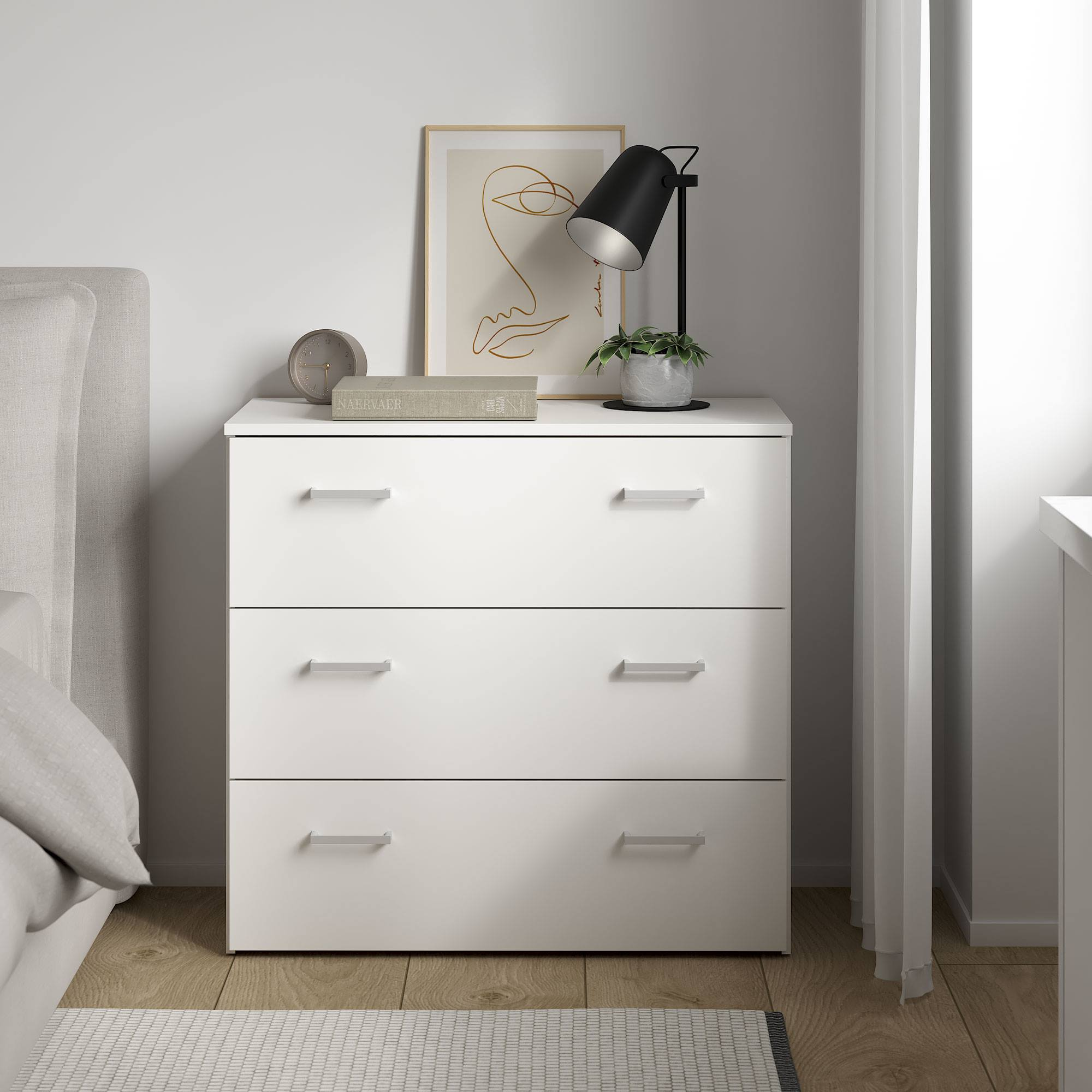 Cómoda blanca con tres cajones. Una lámpara, un cuadro y una planta encima. Una cama es visible a la izquierda.