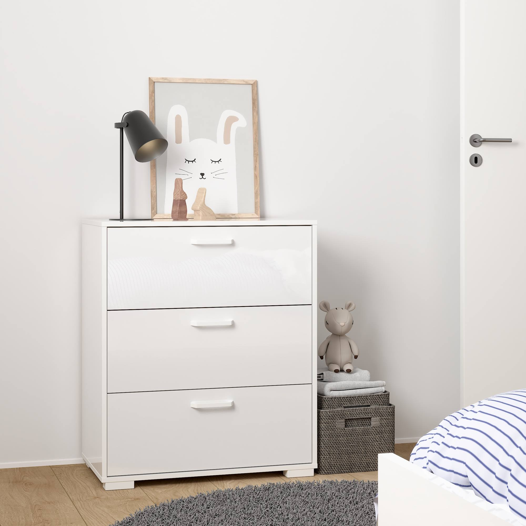 Una cómoda blanca con lámpara y cuadro de conejo. Juguete y cesta al lado. Una cama en la esquina.