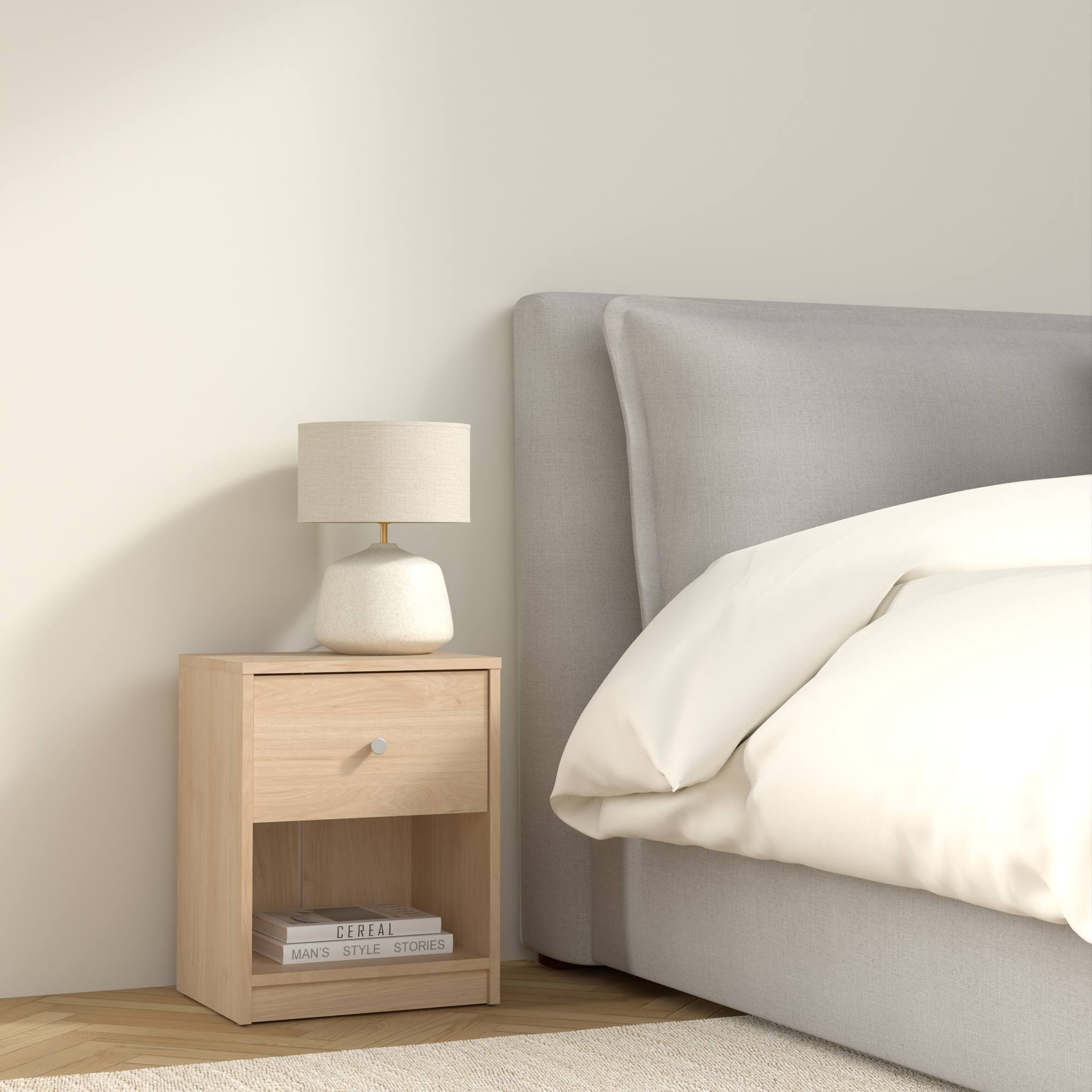 Una mesita de noche con lámpara y libros junto a una cama con ropa de cama blanca. La pared es beige.