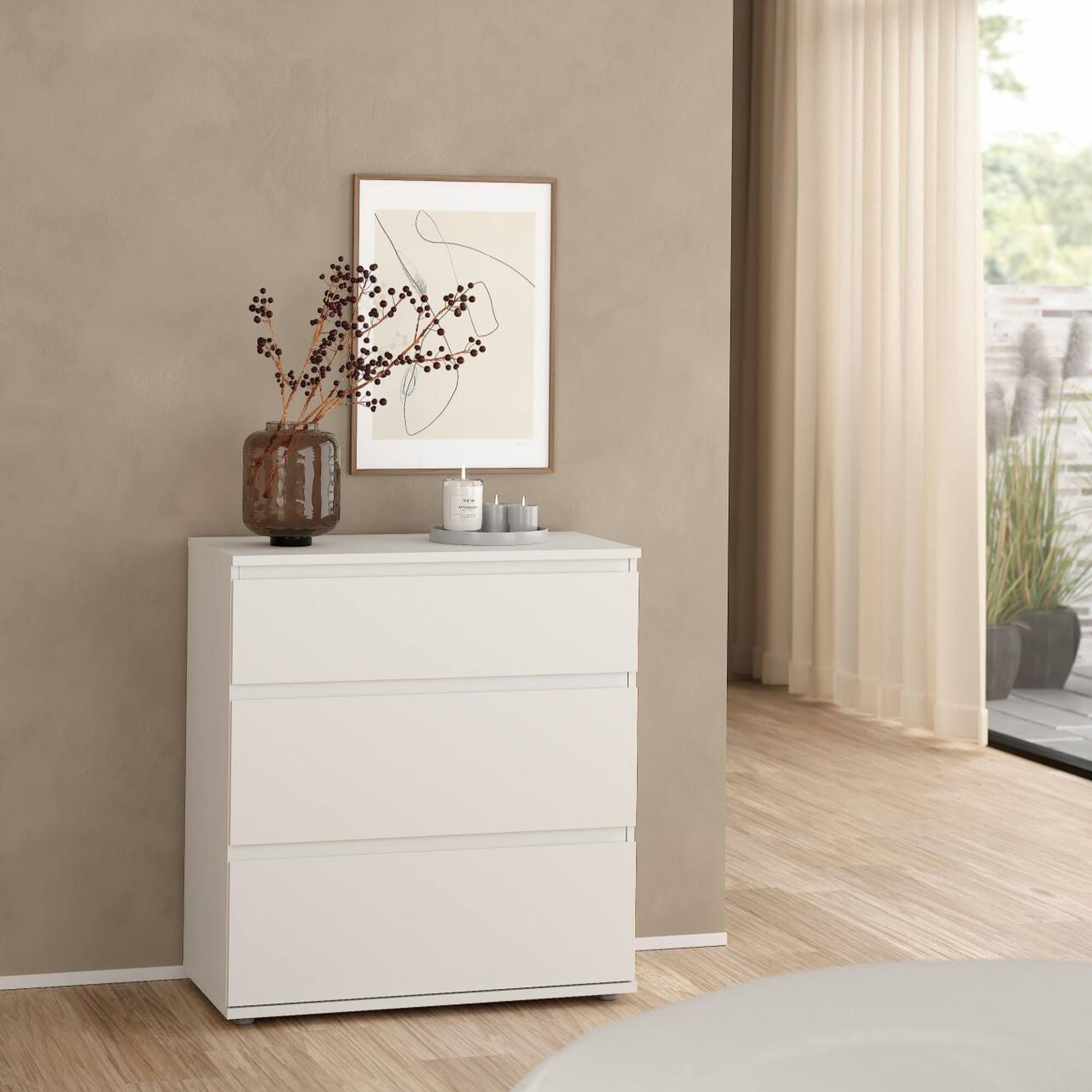 Una cómoda blanca con tres cajones. Encima hay un jarrón con ramas, velas y una obra de arte enmarcada. Hay una ventana en el fondo.