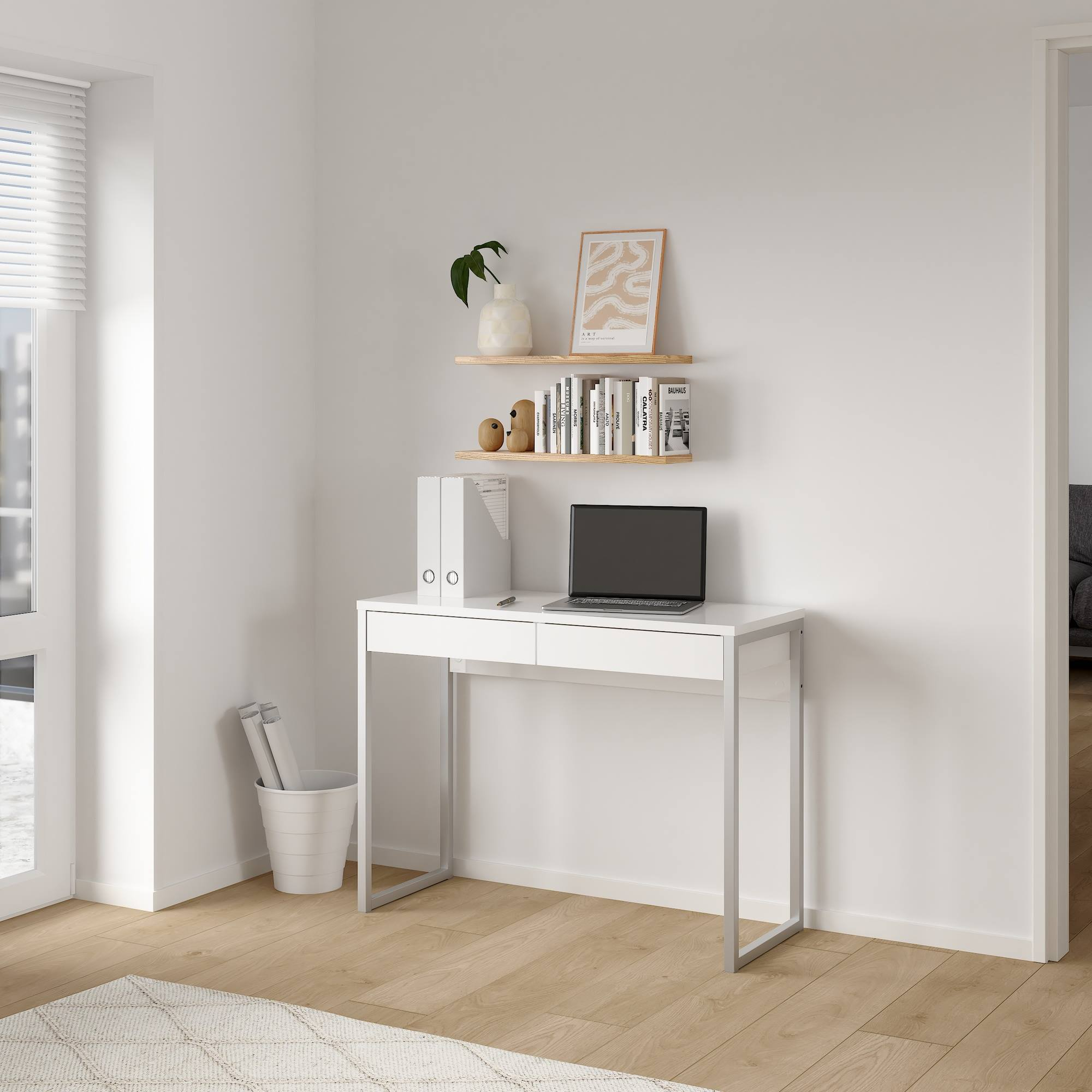Escritorio con laptop, estantes con libros y decoración, paredes blancas, piso de madera, ventana con persianas y alfombra blanca.