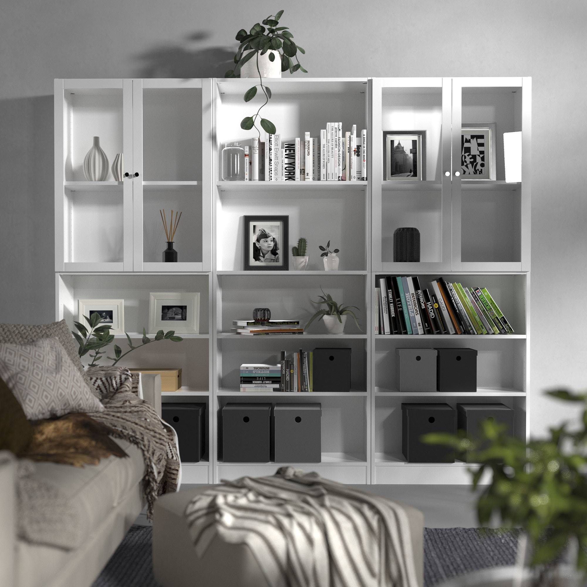 Una estantería blanca con varios objetos: libros, cuadros, plantas y objetos decorativos. Un sofá y un otomano en primer plano.