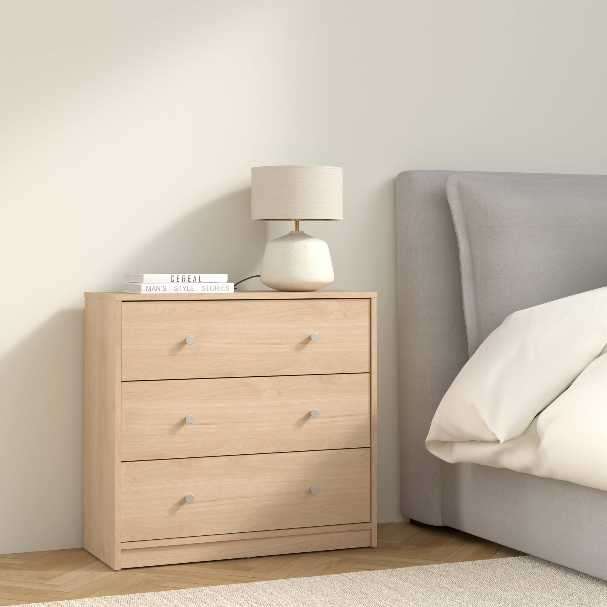Cómoda de madera con lámpara y libros. A la derecha, una cama con cabecero gris.