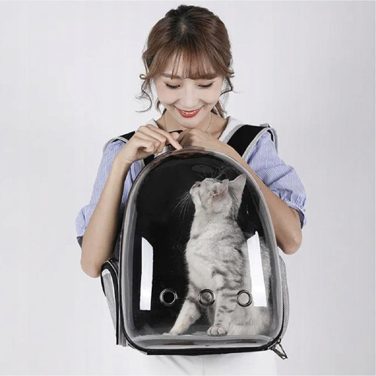 Mujer sosteniendo un gato en una mochila transportadora. El gato es gris y blanco, mirando hacia arriba. El transportador tiene una burbuja transparente.
