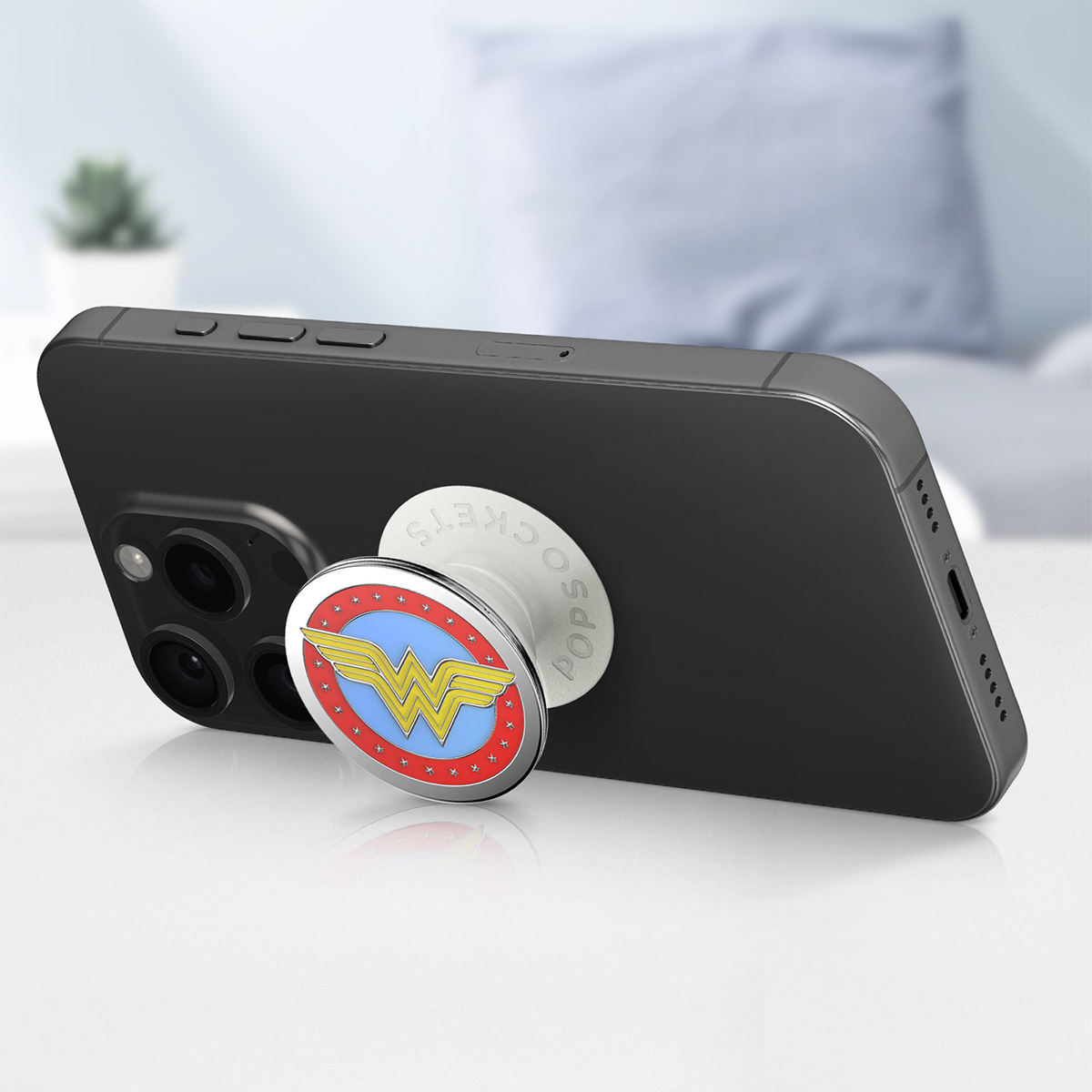 Smartphone nero con PopSocket di Wonder Woman su una superficie bianca. Lo sfondo mostra una stanza sfocata.