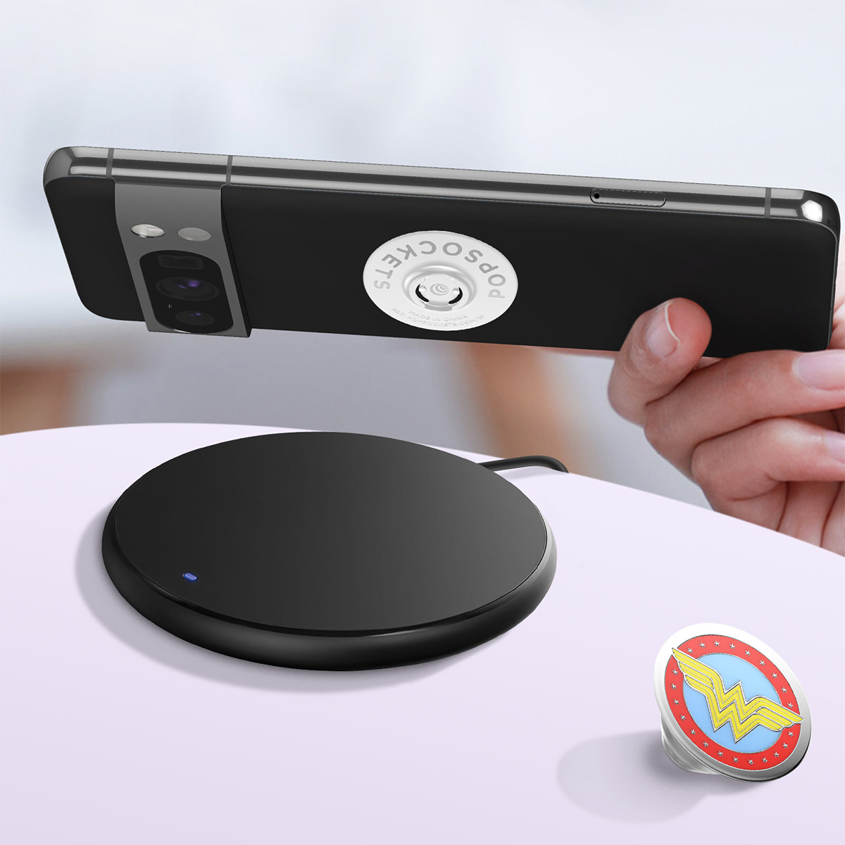Telefono nero con PopSocket, caricatore wireless e PopSocket di Wonder Woman su una superficie bianca. Mano che tiene il telefono.