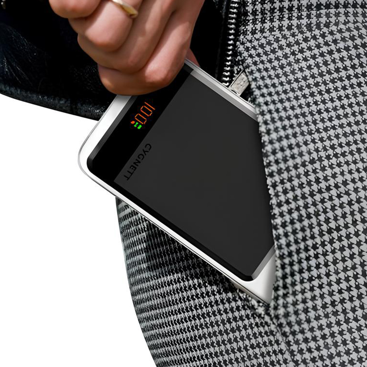 Eine Person hält eine schwarze und silberne Cygnnet Powerbank, die 100 % Ladung anzeigt. Sie befindet sich teilweise in einer Tasche einer Hahnentrittjacke.