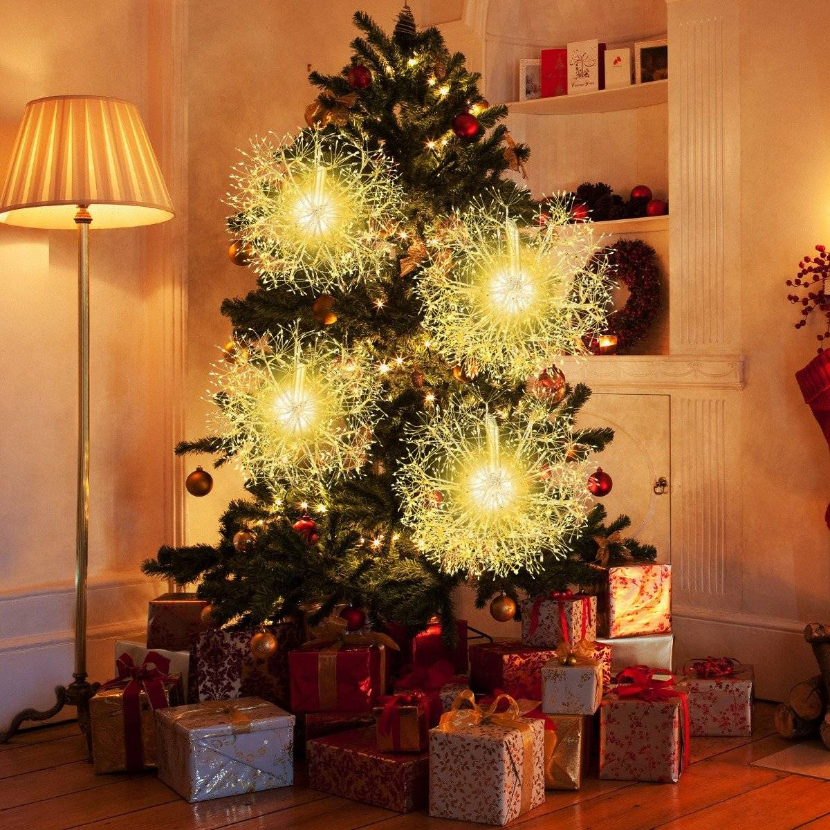 Árbol de Navidad con luces, adornos y regalos. Una lámpara a la izquierda. Una corona y una chimenea al fondo.