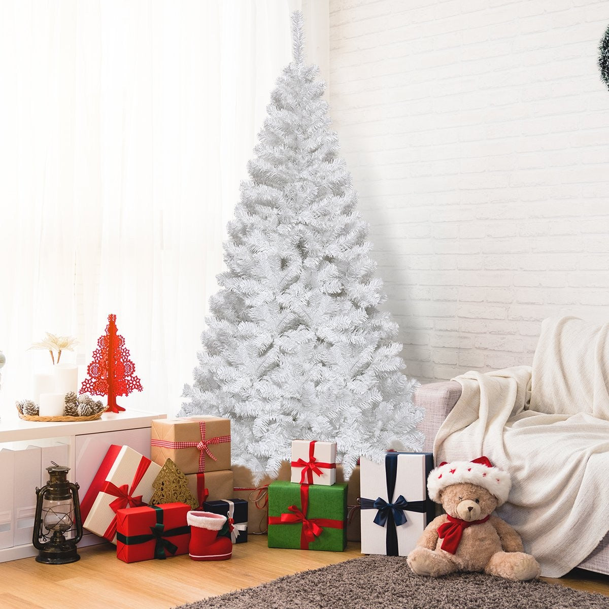 Un árbol de Navidad blanco cerca de regalos envueltos y un oso de peluche con gorro de Papá Noel, junto a un sofá.