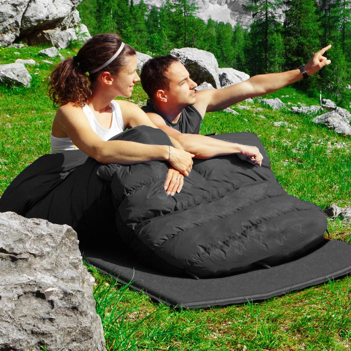 Una pareja sentada en un saco de dormir negro en una zona de césped, señalando un paisaje montañoso con árboles.