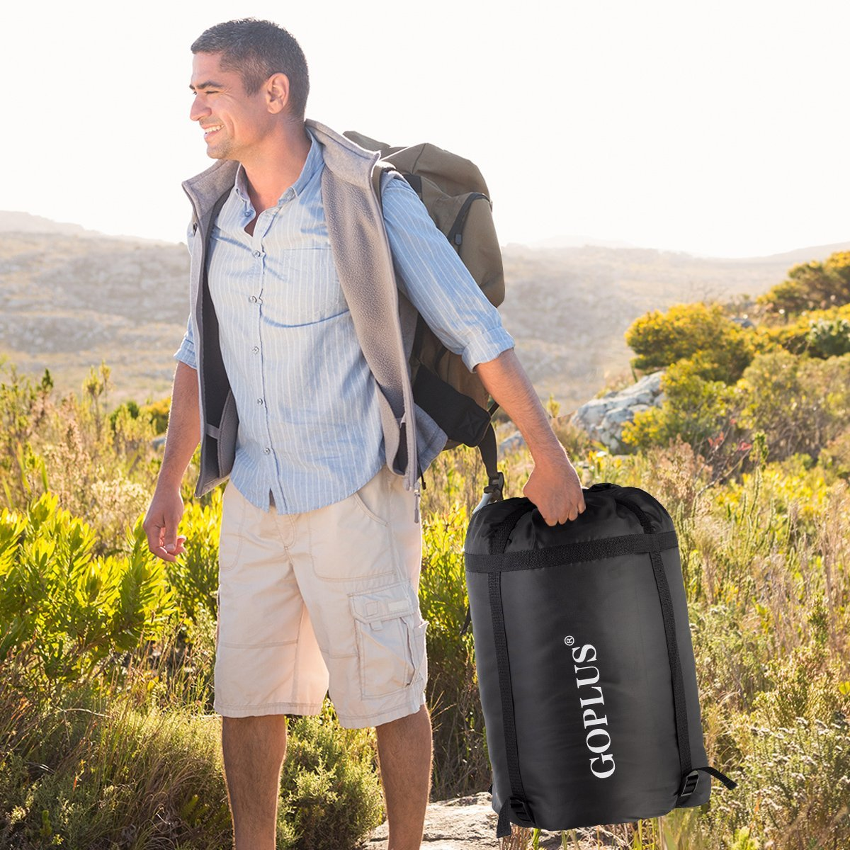 Un hombre camina, llevando una mochila y un saco de dormir negro. Sonríe, caminando por un campo soleado con plantas verdes.