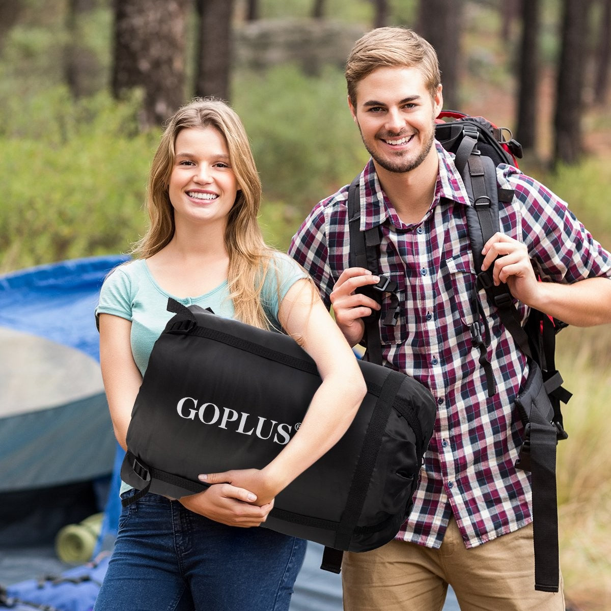 Una pareja sonríe, sosteniendo equipo de campamento en un bosque. Uno lleva una bolsa negra, el otro una mochila.