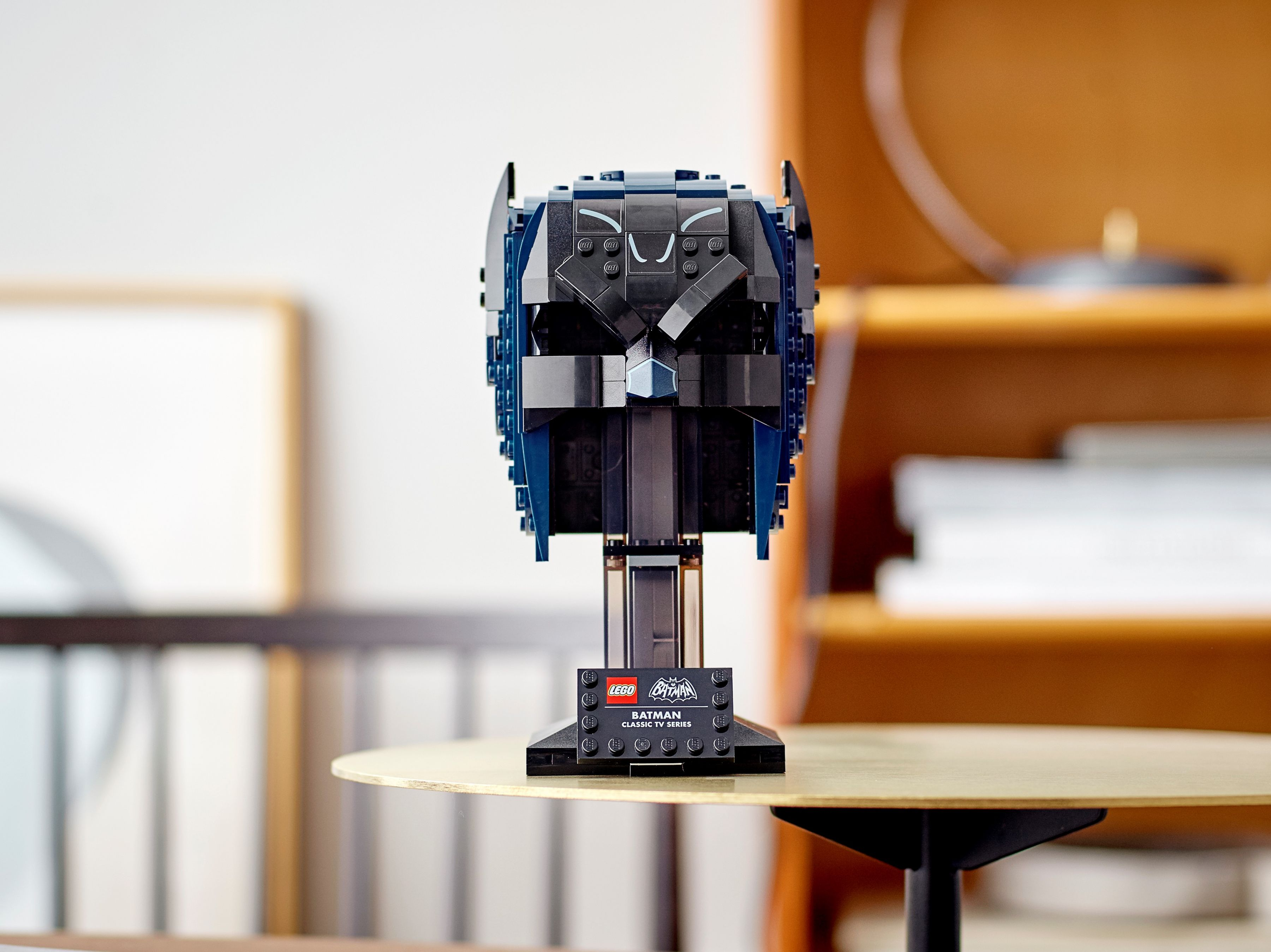 Lego Batman-Helm auf einem Ständer, schwarz und blau, auf einem goldenen Tisch. Hintergrund: Holzregal und gerahmte Kunst.