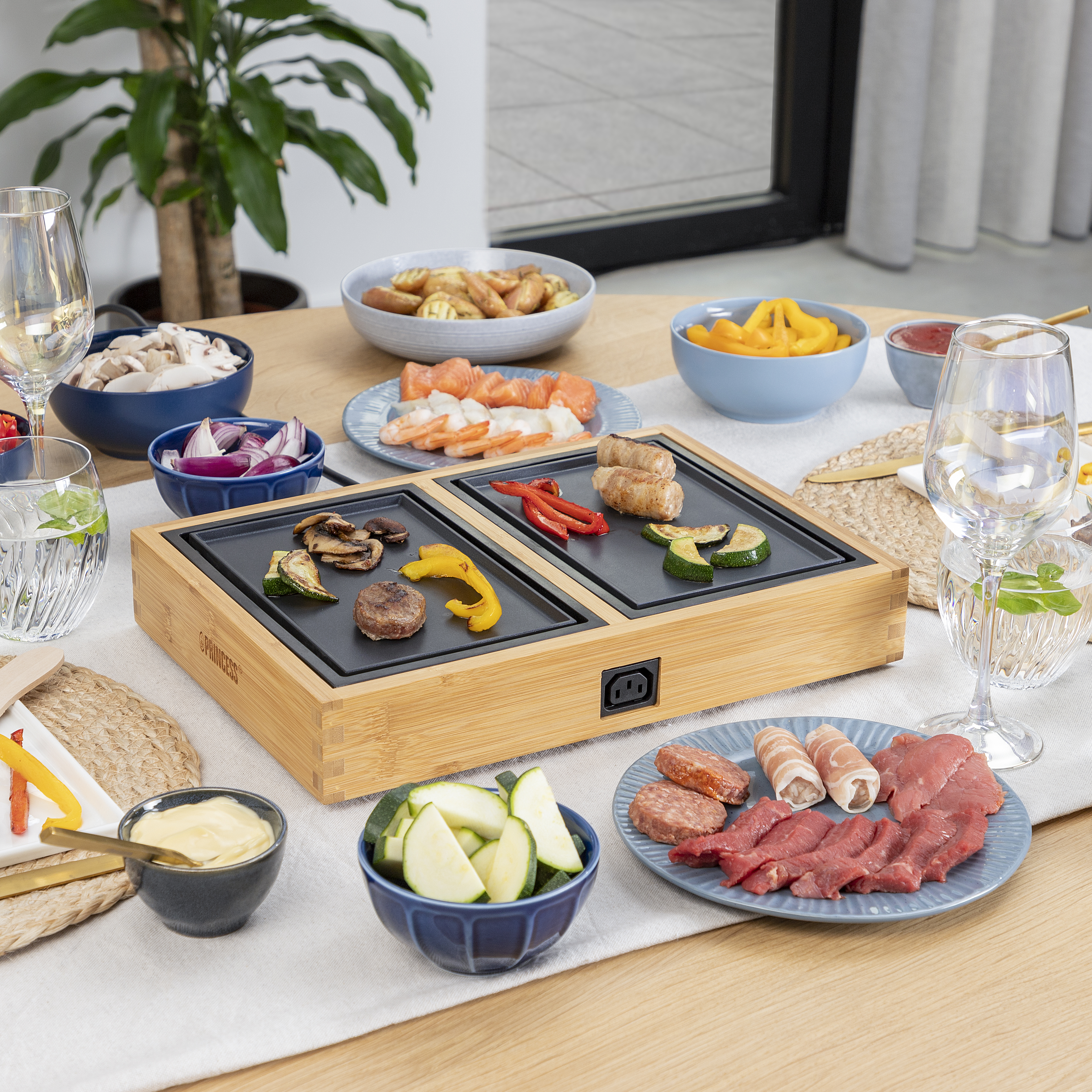 Tisch mit Zutaten und Grill. Gemüse, Fleisch und Getränke. Im Hintergrund eine Pflanze.
