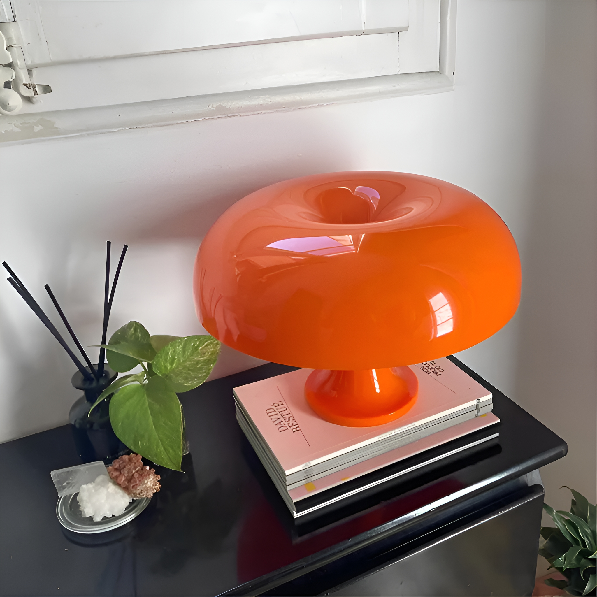 Eine orangefarbene Pilzlampe steht auf Büchern. Ein schwarzer Tisch trägt Pflanzen, Kristalle und einen Diffusor. Weißer Wandhintergrund.
