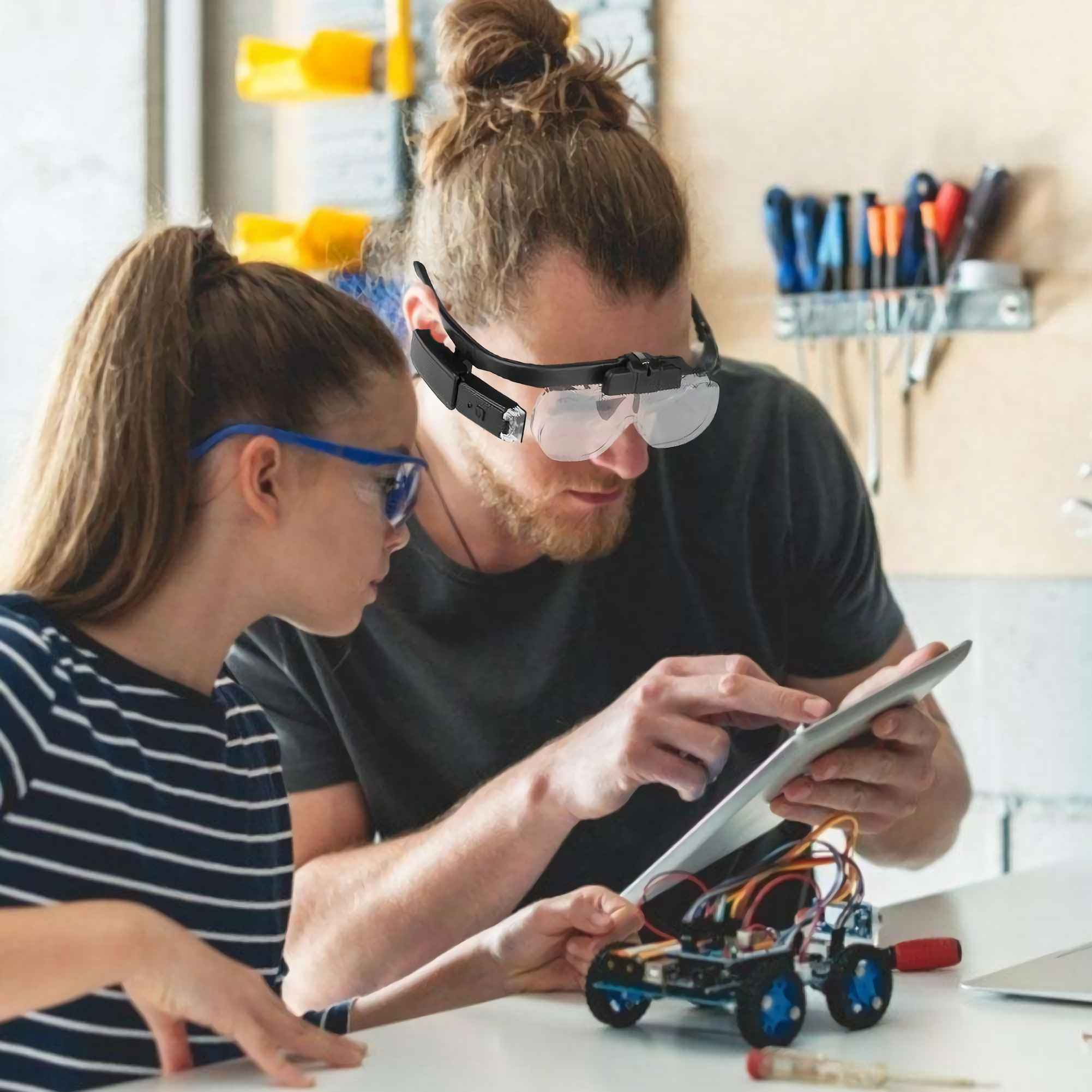 Zwei Personen betrachten ein Tablet und arbeiten an einem Roboter. Einer trägt eine Kopfbandlupe. Werkzeuge sind im Hintergrund.