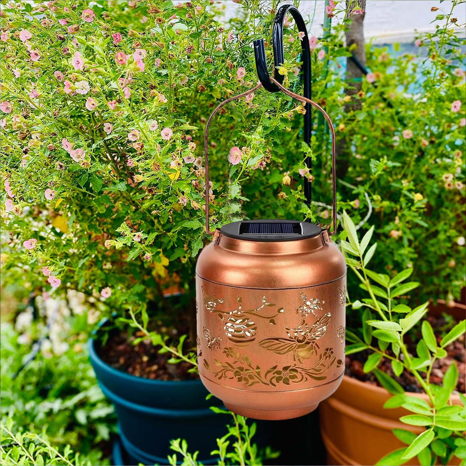 Eine Kupferlaterne mit floralen Ausschnitten hängt an einem schwarzen Haken in einem Garten mit Pflanzen und rosa Blumen.