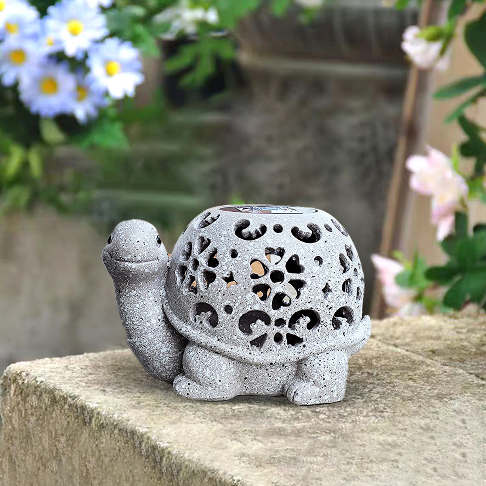 Eine graue Schildkrötenstatue mit floralen Ausschnitten sitzt auf einem Steinsims. Blumen sind im Hintergrund.