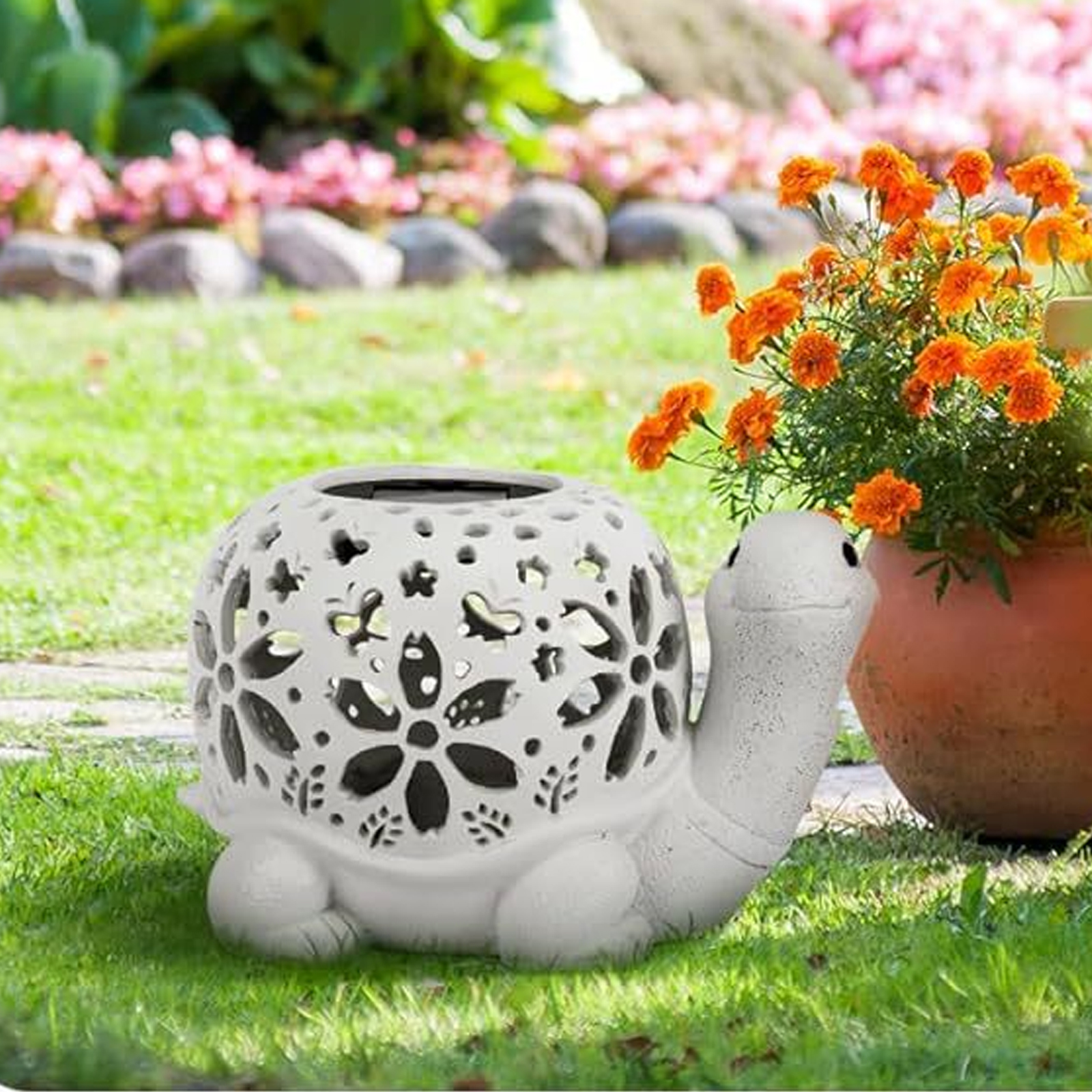 Ein weißes, schildkrötenförmiges Gartenornament mit floralen Ausschnitten. In der Nähe befinden sich orangefarbene Blumen in einem Terrakottatopf auf dem Rasen.