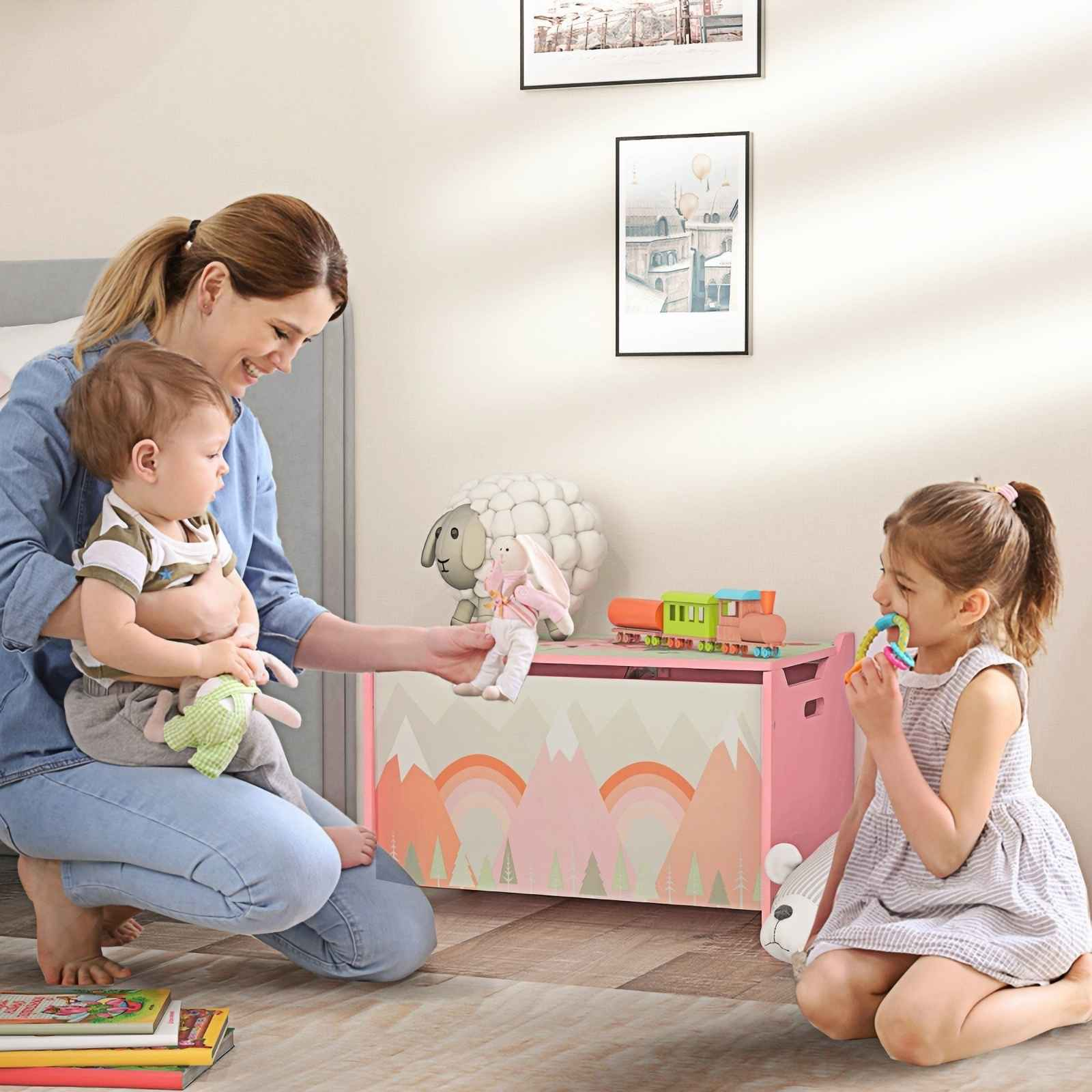 Eine Frau hält ein Baby, ein Mädchen kaut an einem Spielzeug, eine Spielzeugkiste mit Bergen und ein Schafspielzeug im Raum.