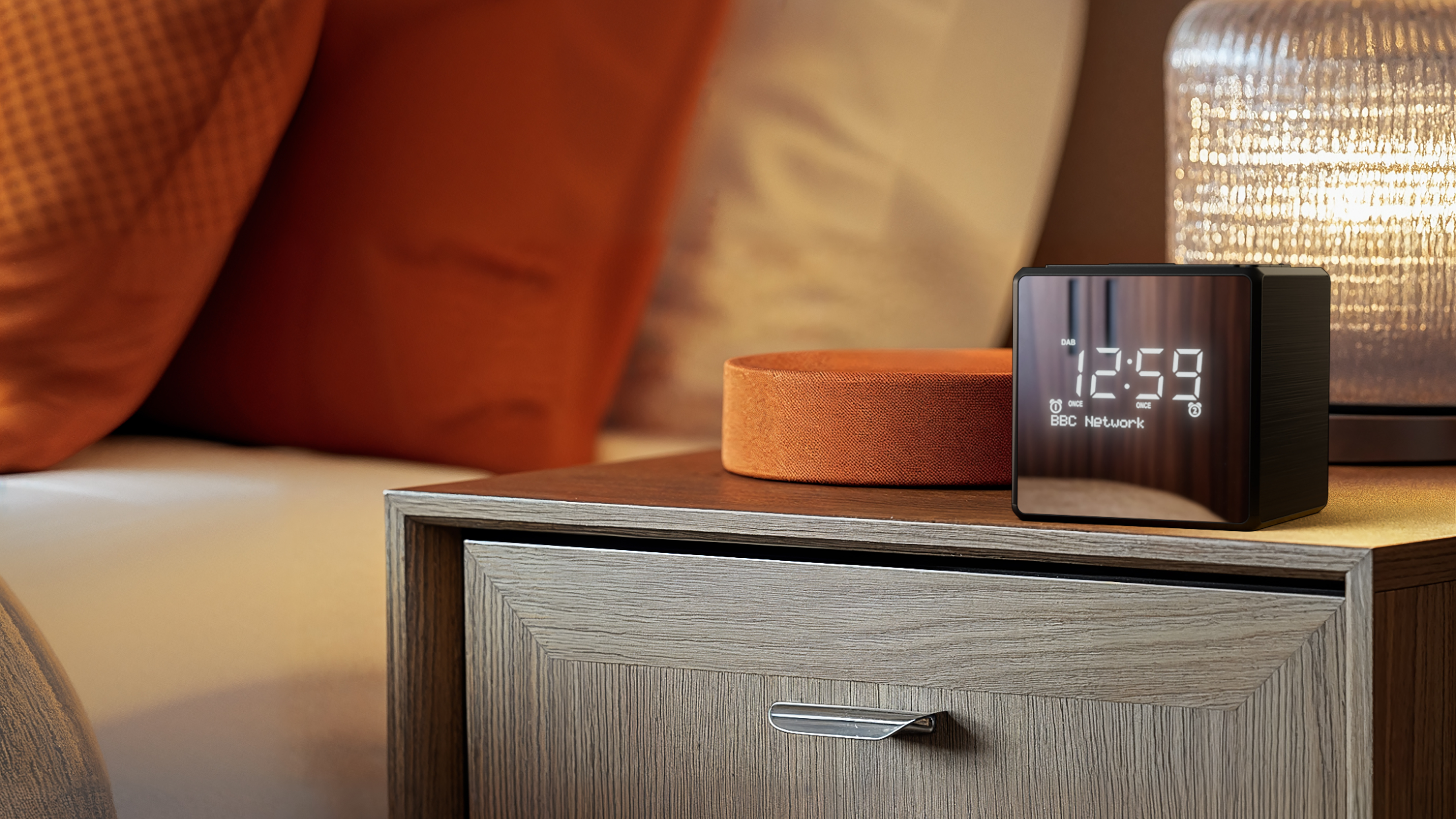 Ein Nachttisch mit Uhr (12:59), einem kreisförmigen Objekt und einer Lampe. Orangefarbene Kissen im Hintergrund.