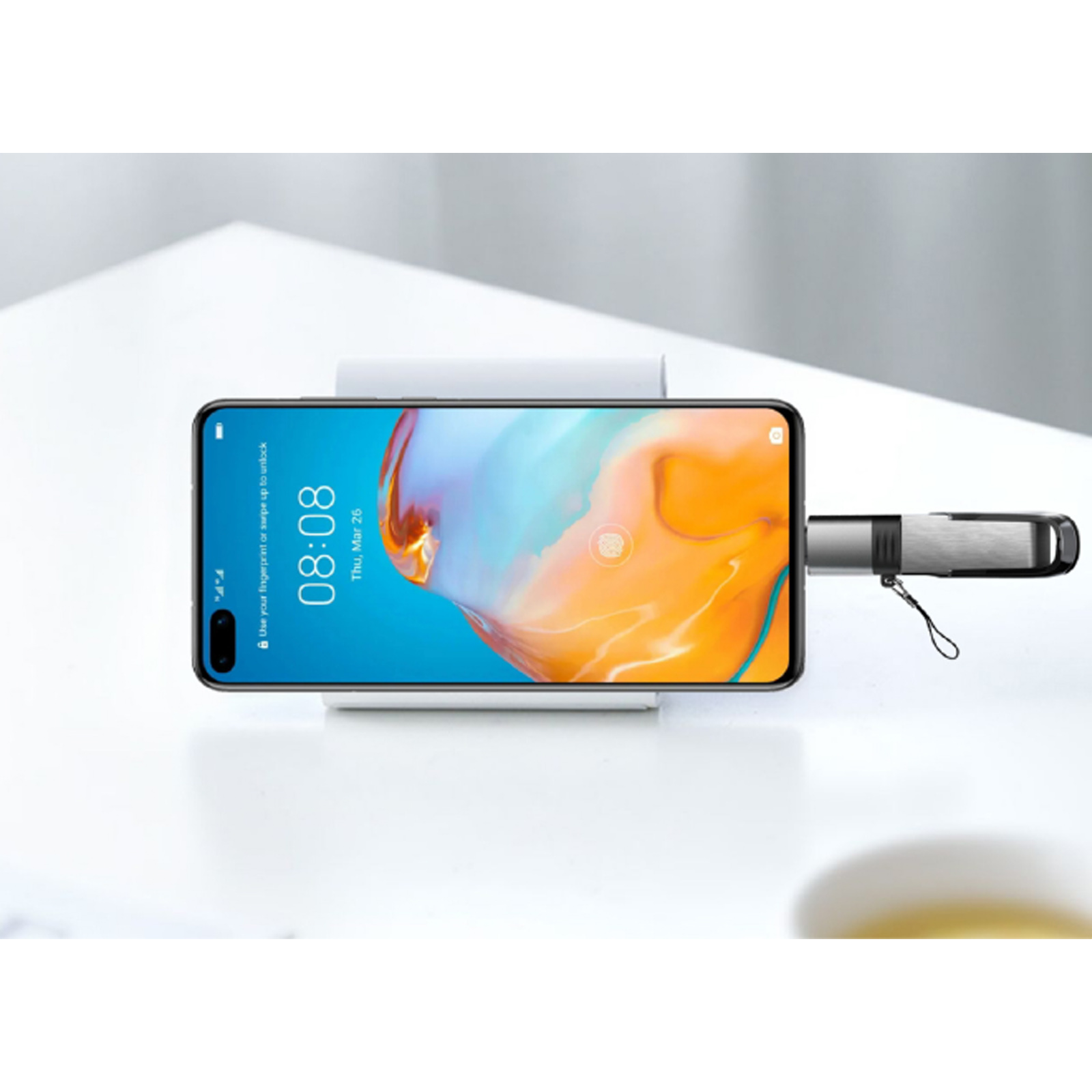 Un smartphone en un soporte blanco muestra 08:08. Una unidad USB-C está conectada. Superficie blanca de fondo.