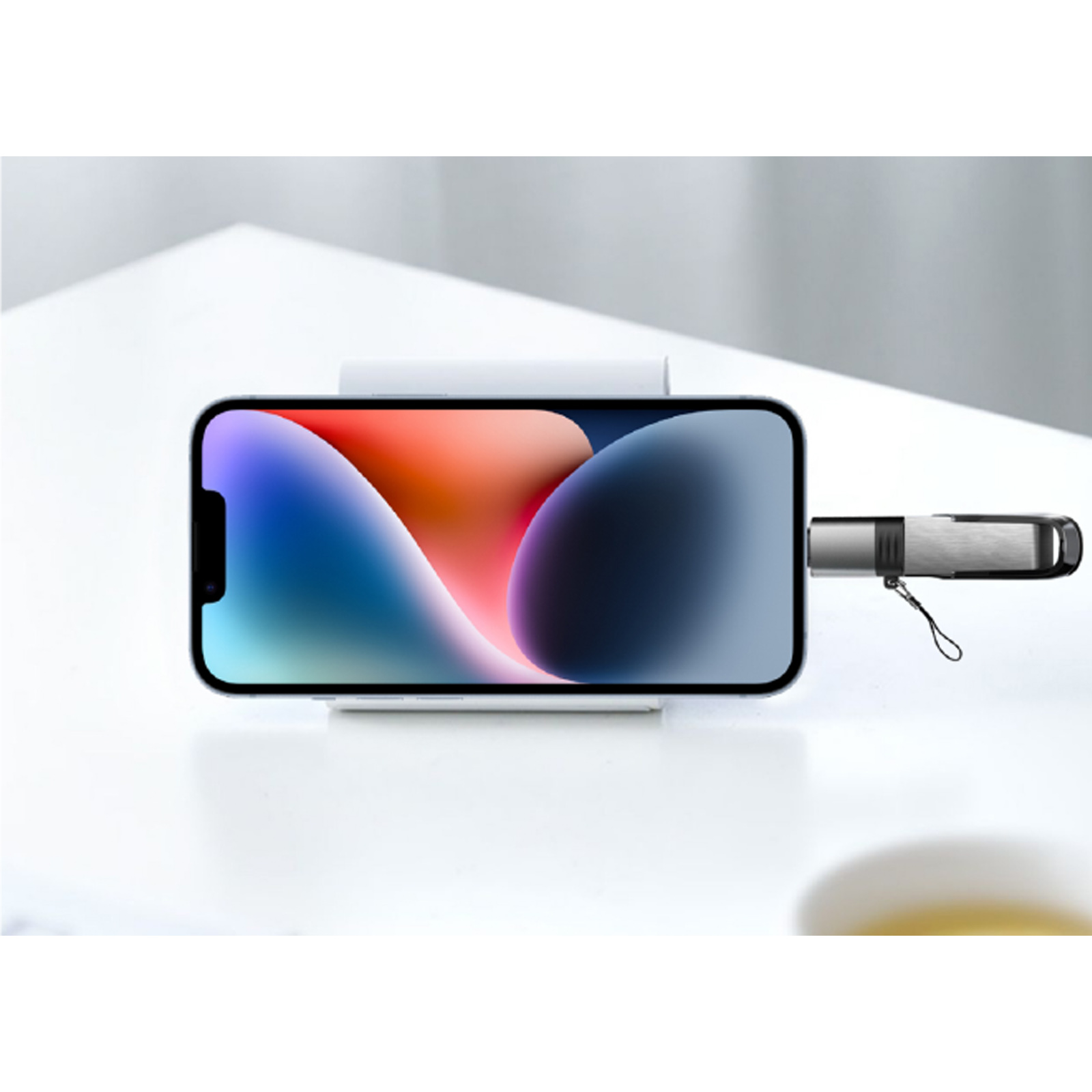 Un iPhone descansa sobre una base de carga blanca. Hay una unidad USB conectada. La pantalla del teléfono muestra un fondo colorido.