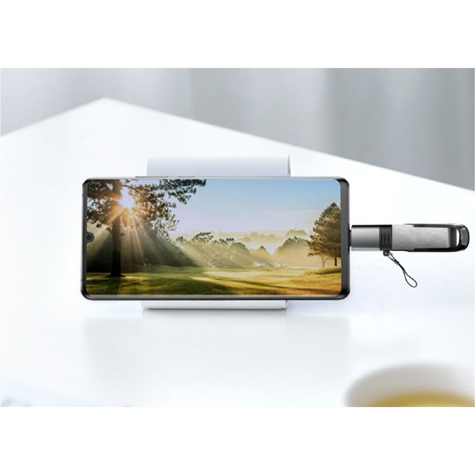 Un teléfono muestra una foto de paisaje, conectado a una unidad USB. La escena está sobre una superficie blanca.