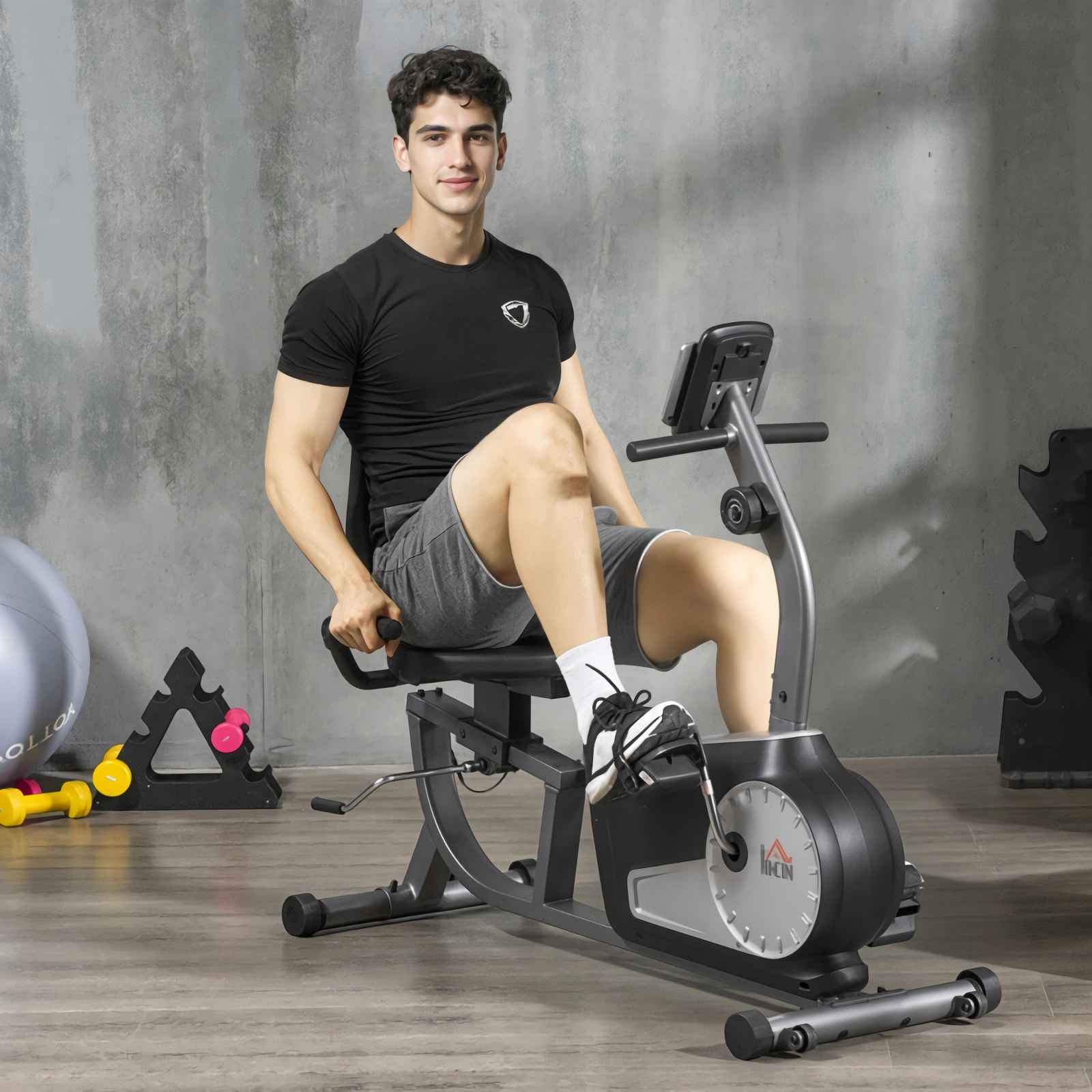 Mann auf Liegefahrrad. Er trägt ein schwarzes Hemd, graue Shorts und weiße Socken. Fitnessgeräte sind im Hintergrund.