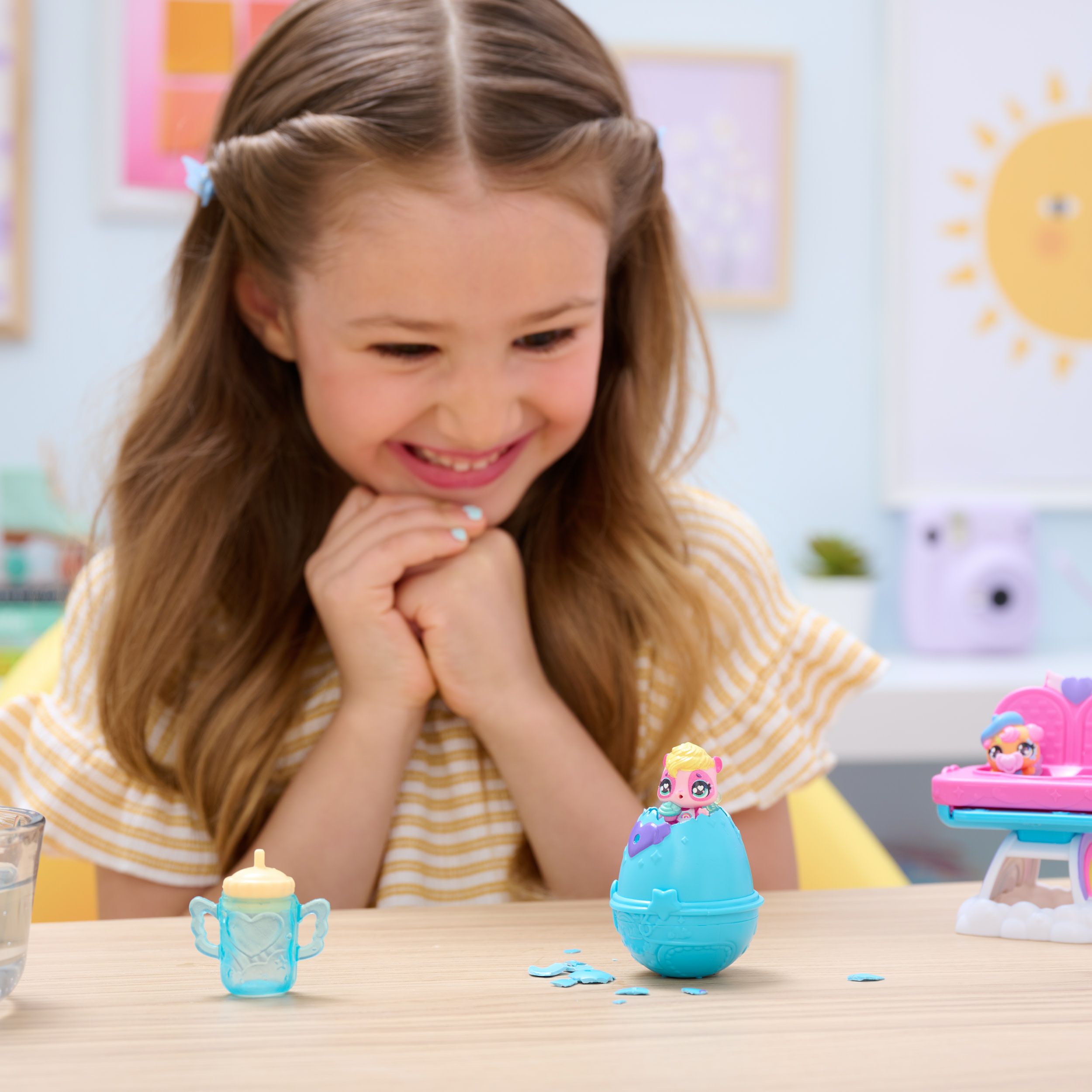 Ein Mädchen lächelt Spielzeug auf einem Tisch an. Enthält eine Babyflasche, ein Spielzeug-Ei und ein Spielzeugauto. Der Hintergrund hat eine Sonnenzeichnung.