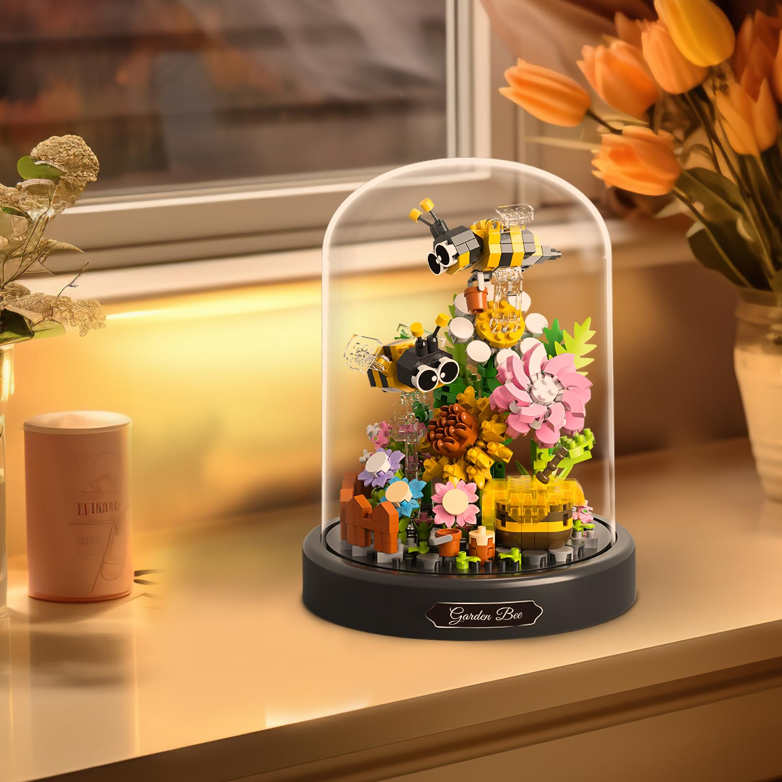 Ein Lego-Bienengarten in einer Glaskuppel. Mit Bienen, Blumen und einem Bienenstock. Auf einer Fensterbank mit Tulpen im Hintergrund.