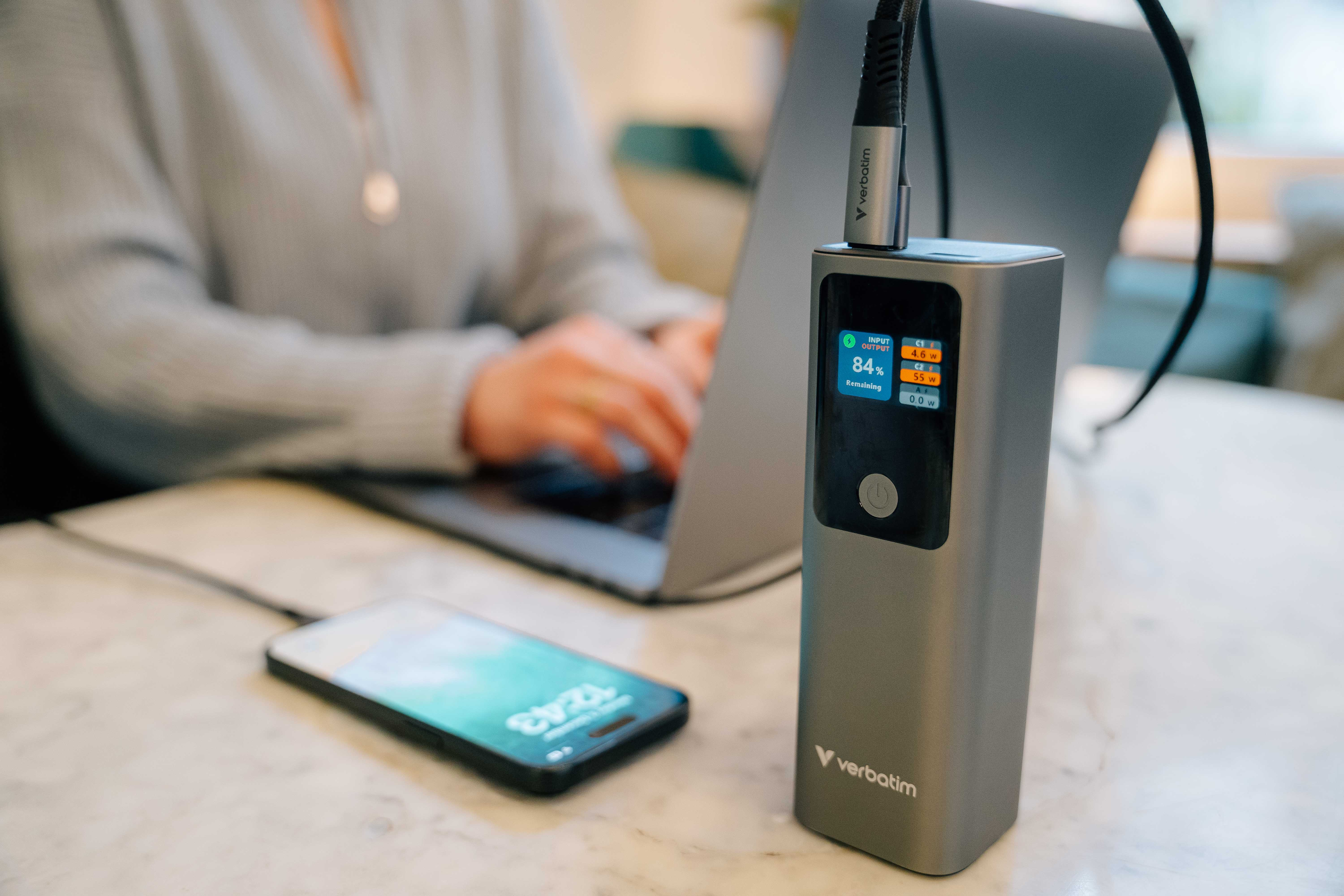 Een persoon gebruikt een laptop. Een powerbank met display toont batterij en een telefoon wordt opgeladen.