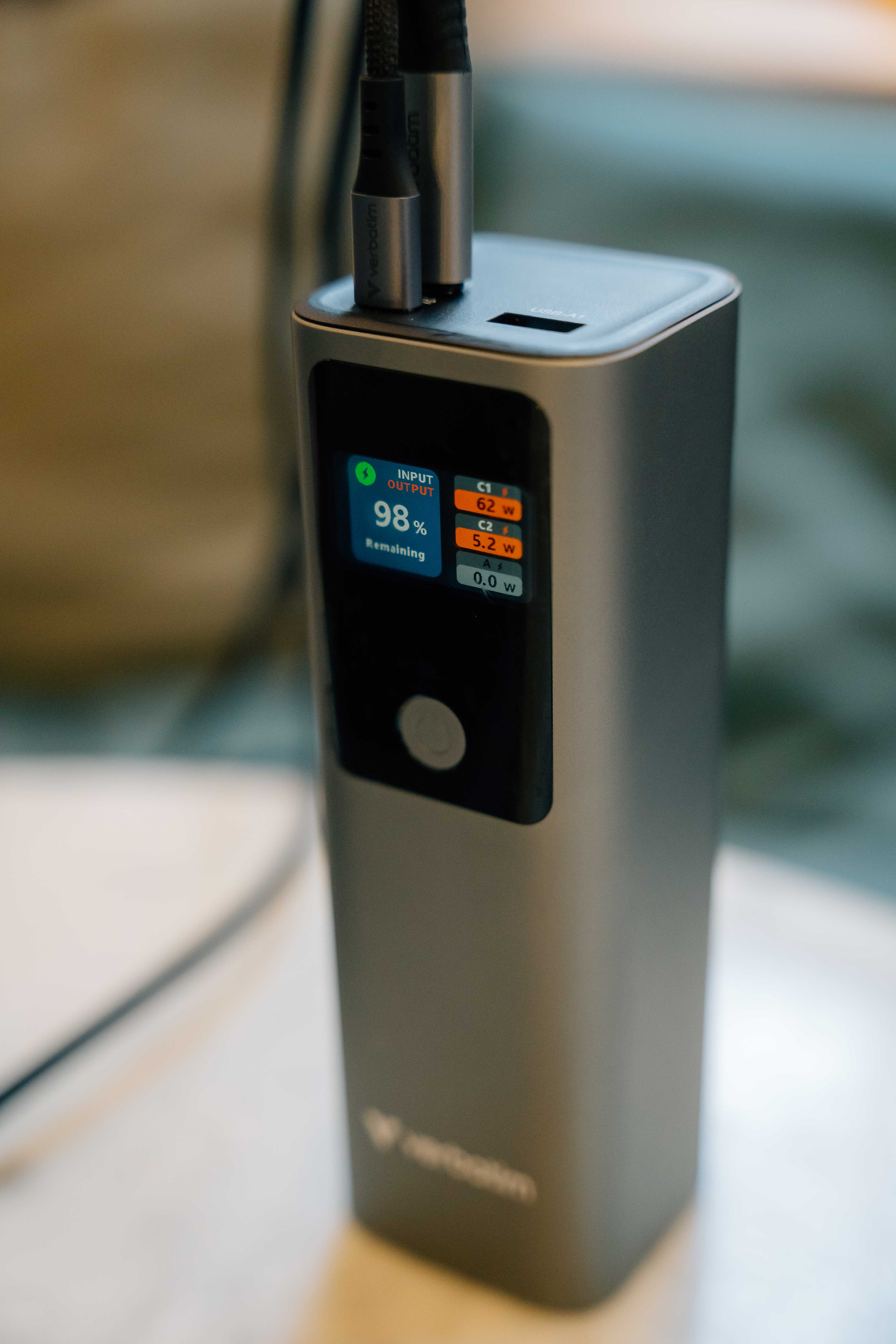 Een zilveren powerbank met een scherm dat de batterijduur en de output weergeeft. Twee zwarte kabels zijn aangesloten.