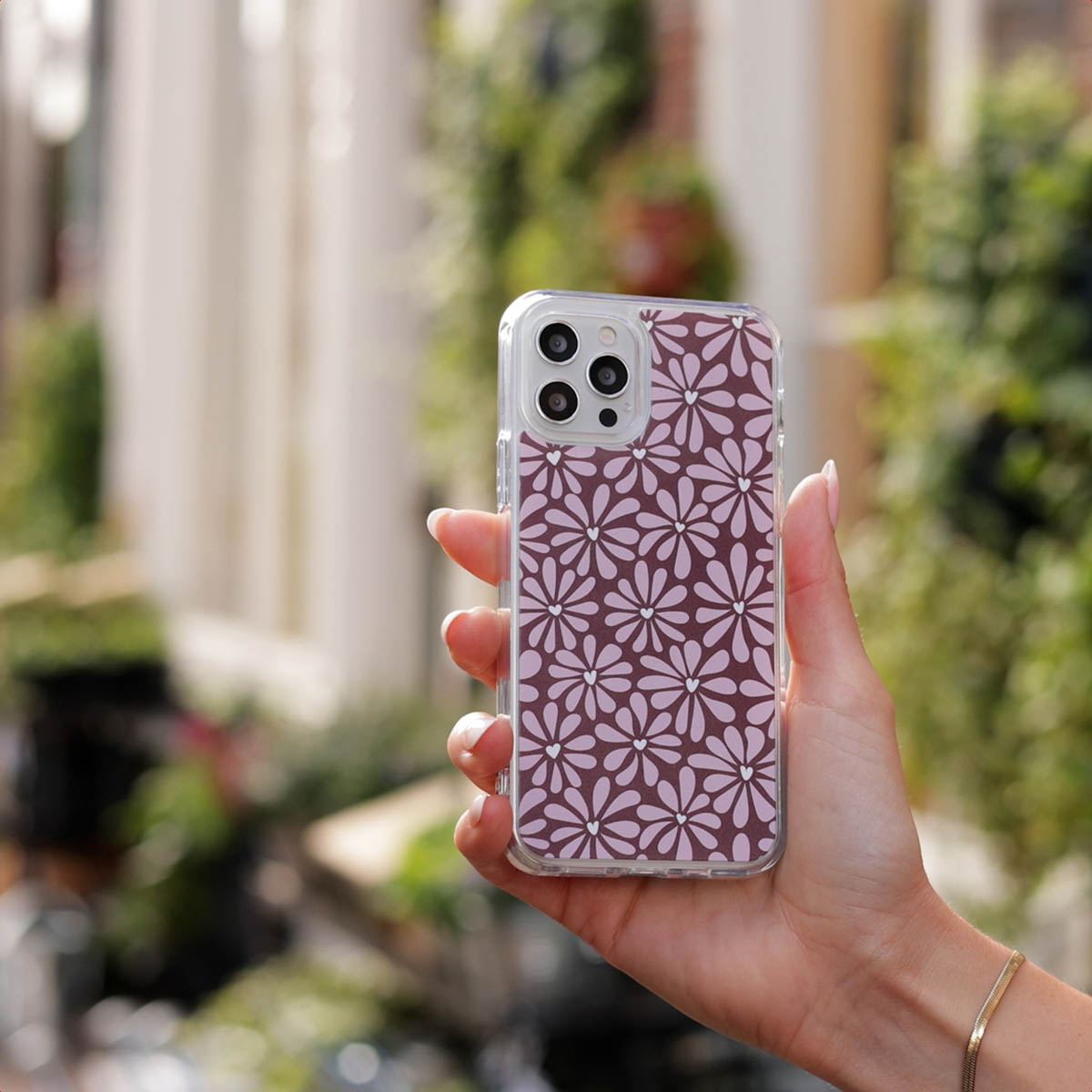 Een hand die een telefoon vasthoudt. De telefoonhoes heeft een bloemenpatroon. De achtergrond is wazig groen gebladerte.