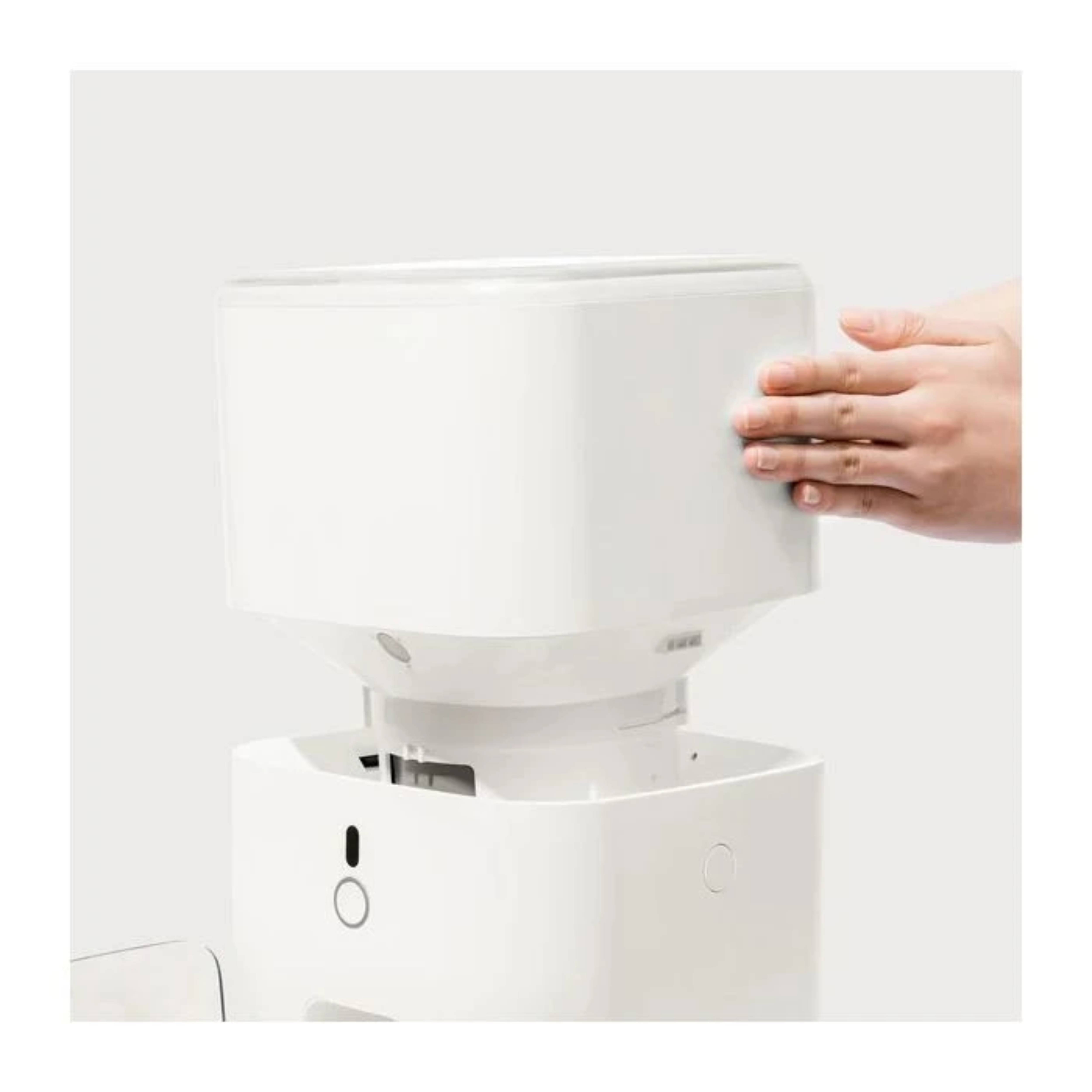 A white pet food dispenser is shown. A hand is touching the top section. The dispenser has a modern design.