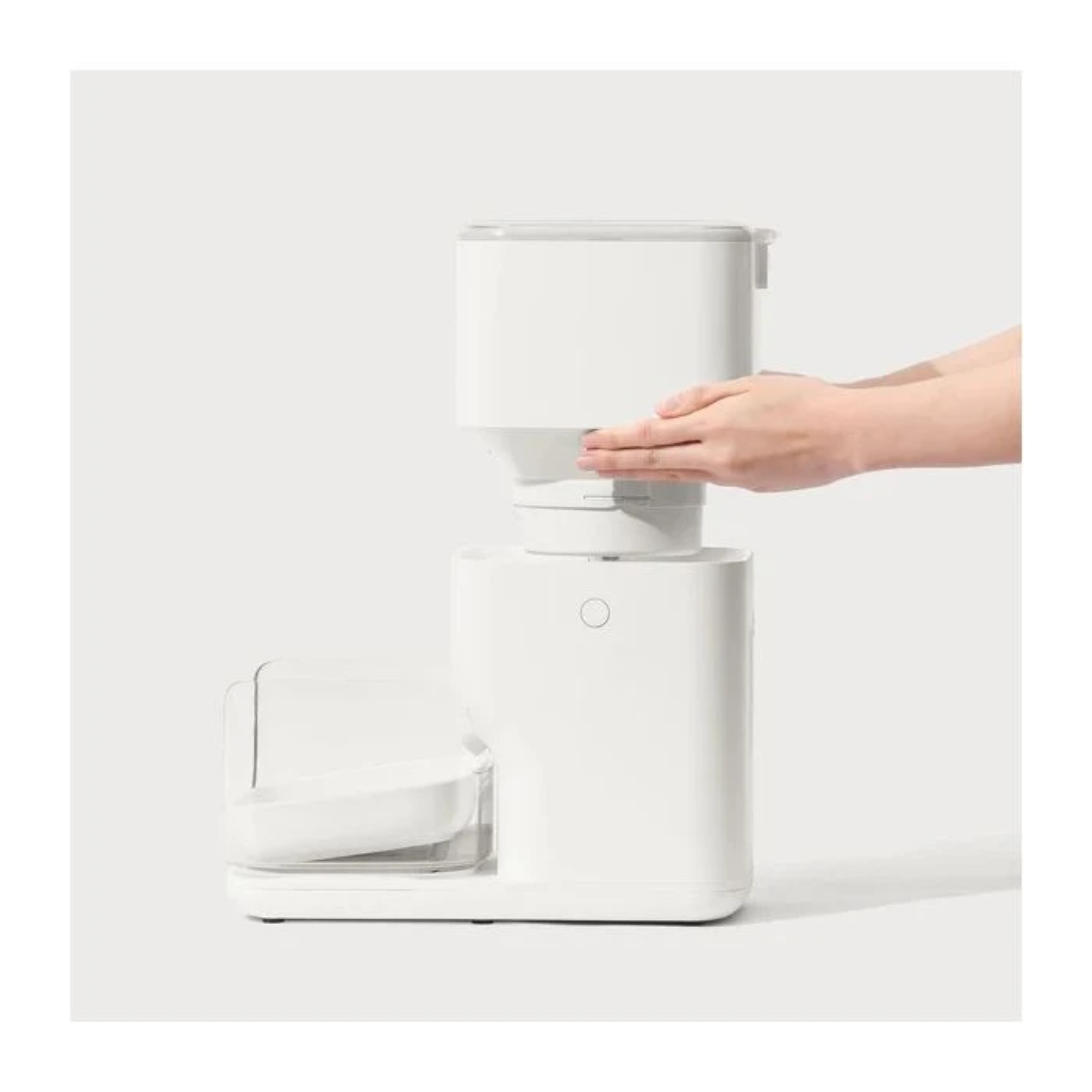A white pet food dispenser. A hand is placing a part on top. A dish is on the side, on a white surface.