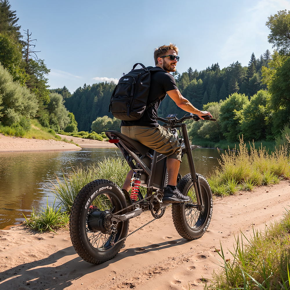 Un hombre monta una bicicleta eléctrica en un camino arenoso junto a un río. La bicicleta tiene neumáticos grandes y el hombre lleva gafas de sol y una mochila.