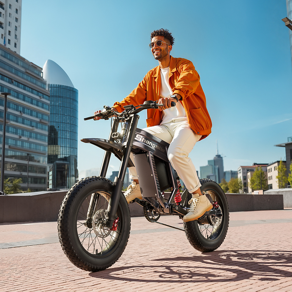 Hombre montando una bicicleta eléctrica, con chaqueta naranja y gafas de sol. Fondo urbano con edificios y cielo azul.