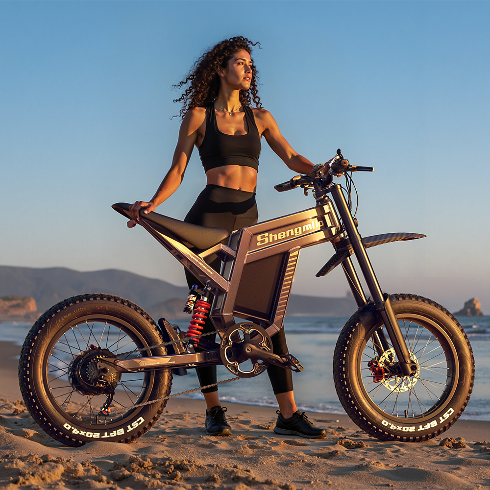 Eine Frau mit lockigem Haar steht neben einem E-Bike am Strand. Sie trägt ein schwarzes Oberteil und Leggings. Das Fahrrad ist grau mit großen Reifen.