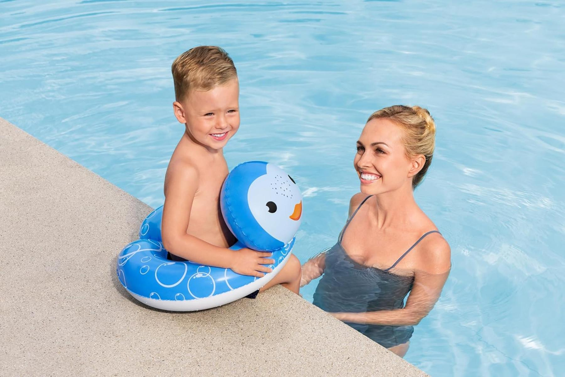 Un niño con un flotador de pingüino azul sonríe a una mujer en una piscina. La mujer también sonríe.