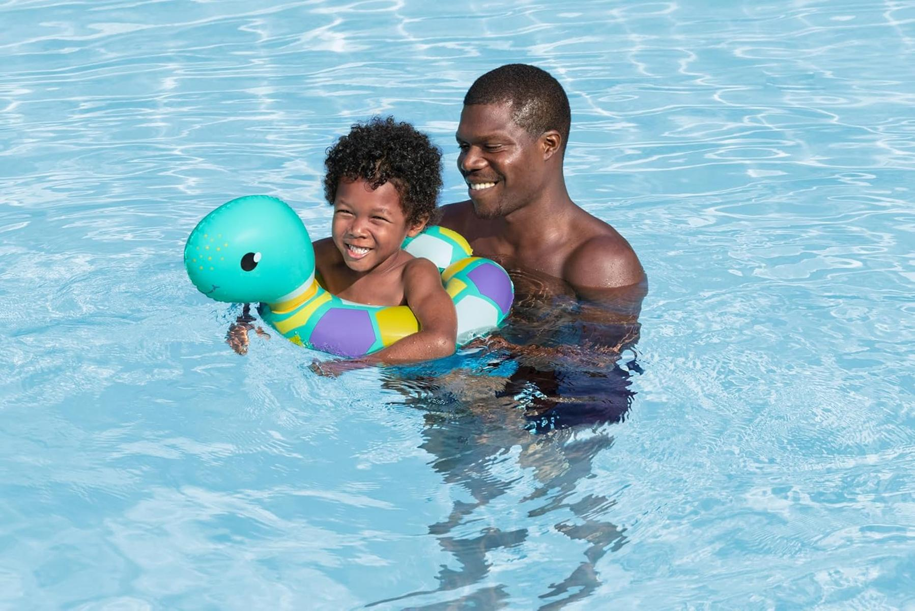 Un hombre y un niño están en una piscina. El niño está en un flotador de tortuga. Ambos están sonriendo. El agua es azul.